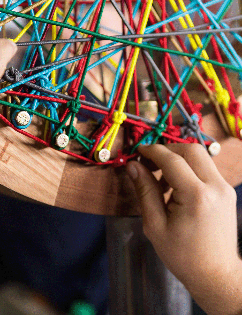 A person's hand weaving a 3D geometric structure using multicolored rubber bands and metal screws on a round wooden board.