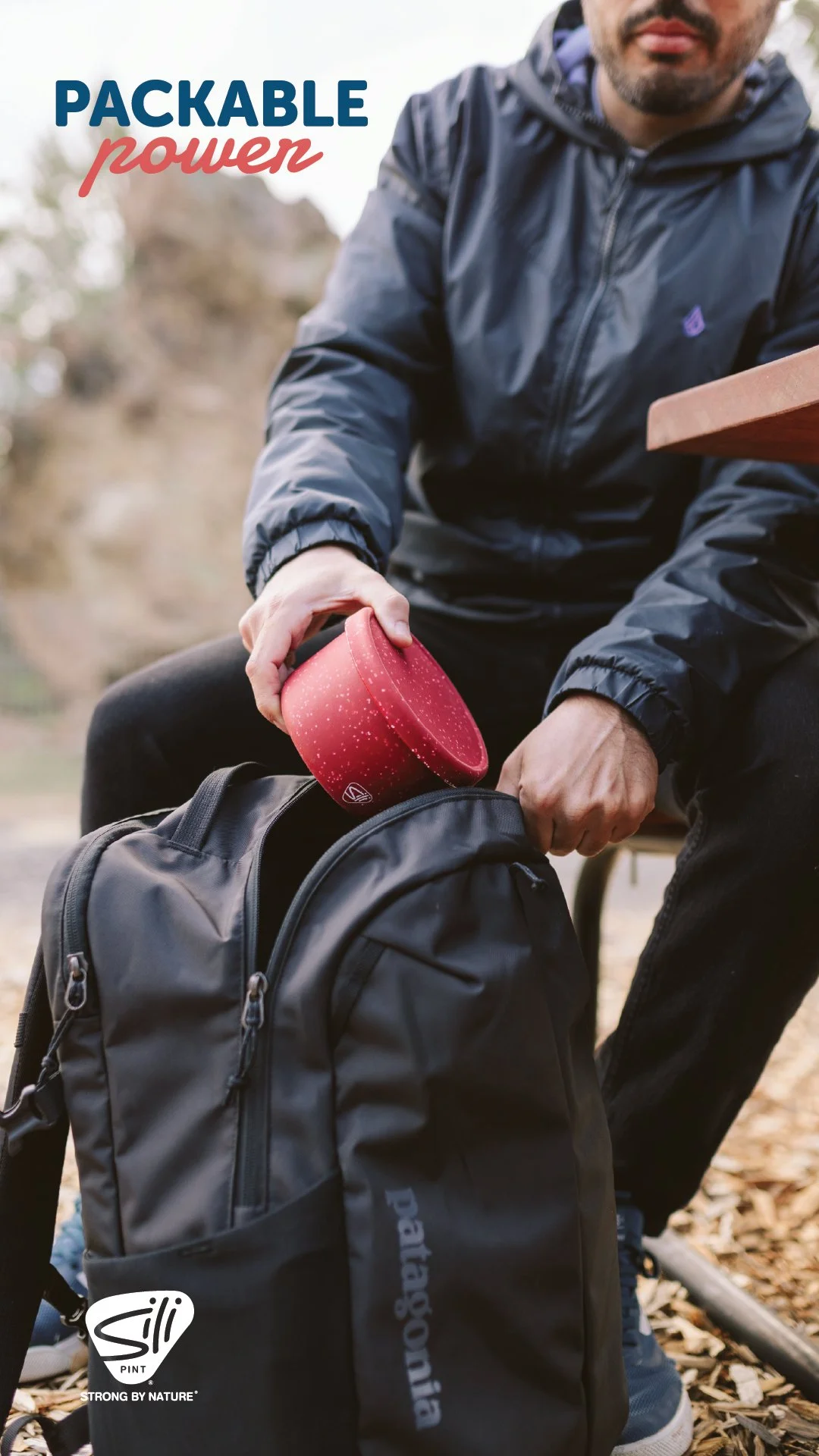 A man in a blue jacket is packing a red portable food container into a black Patagonia backpack outdoors on a trail with wood chips.