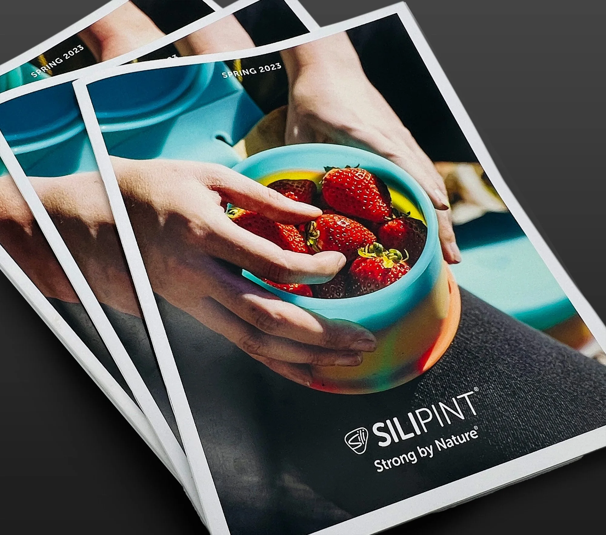 Photograph of a hand holding a colorful silicone bowl filled with strawberries, with a blue silicone bowl in the background, on a blue surface. The text reads 'SPRING 2023 SILIPINT Strong by Nature'.