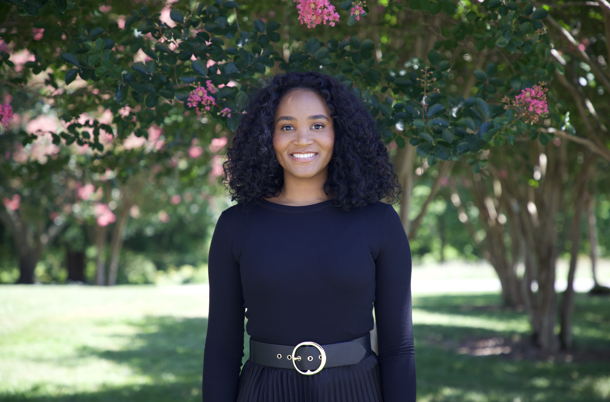 A woman with dark curly hair and brown skin smiling outdoors in front of a flowering tree with pink blossoms and green leaves, wearing a black long-sleeve top and a black pleated skirt with a belt.