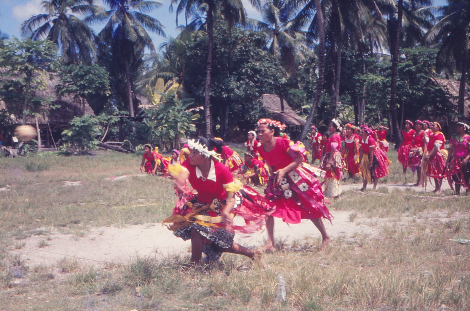 Women dressed in traditional colorful attire playing Ano ball game, with tall palm trees and thatched houses in the background.