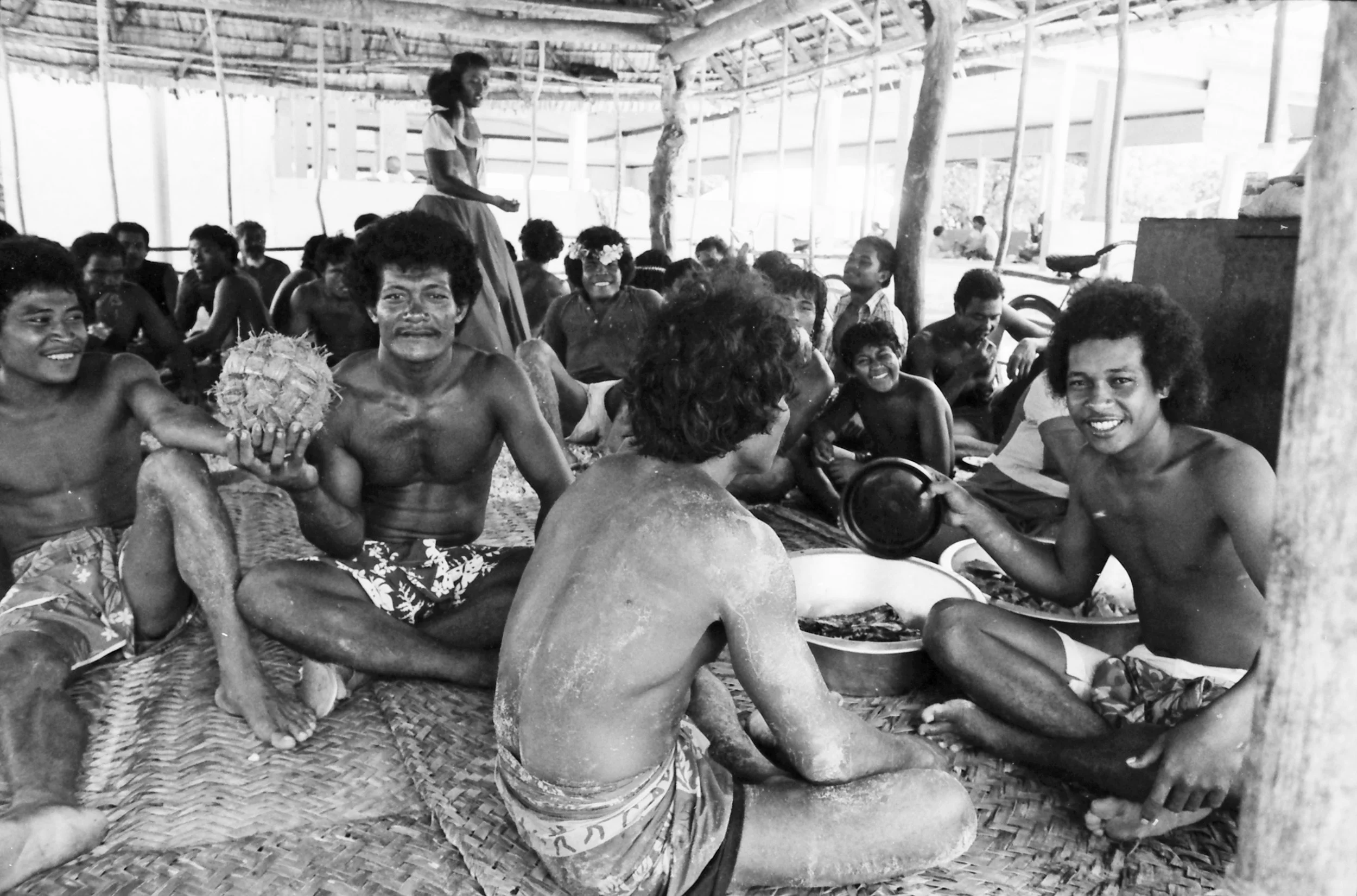 Young men Ano players sitting inside and open-air structure waiting for lunch.  Some people are shirtless and others wearing traditional skirts, smiling and happy, with a woman standing in the background.