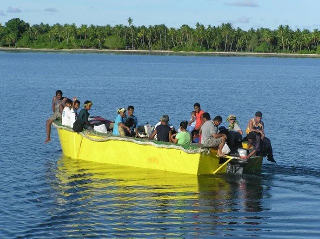 A yellow work boat full of young people on Nanumea's lagoon, heading toward the pass and out to the ship.  Lagoon's far short with with palm trees and greenery in the background.