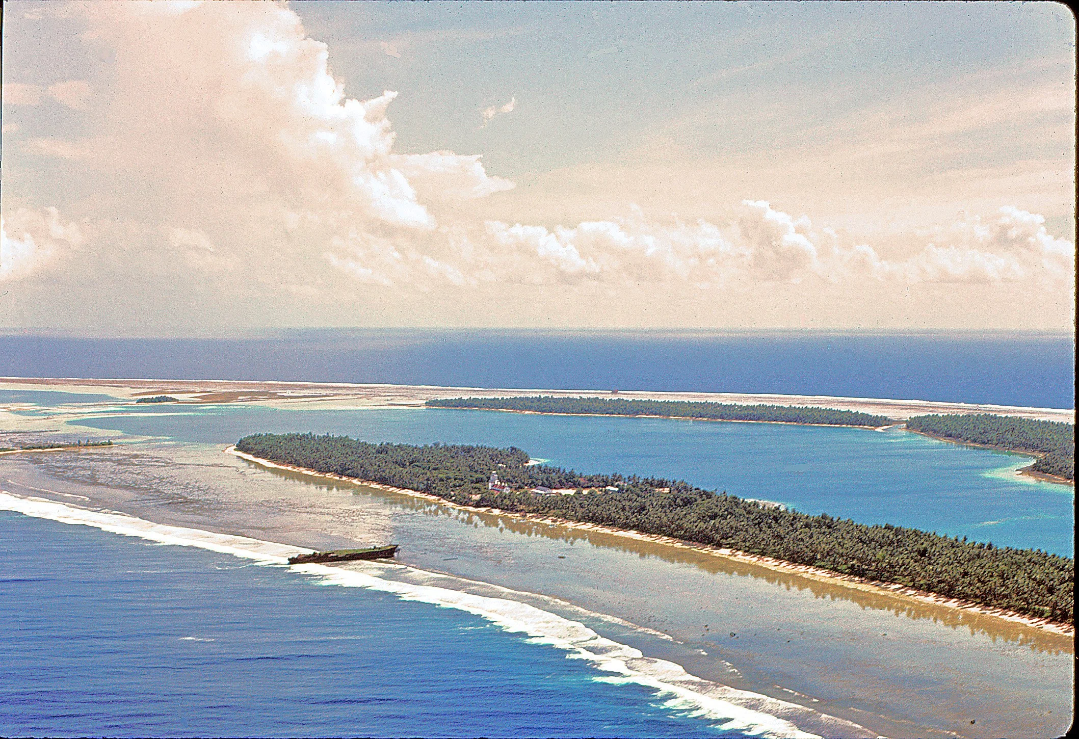 Aerial view of Nanumea island showing village area and lagoon turquoise waters and a barrier reef. The open ocean is visible in the background under a partly cloudy sky.