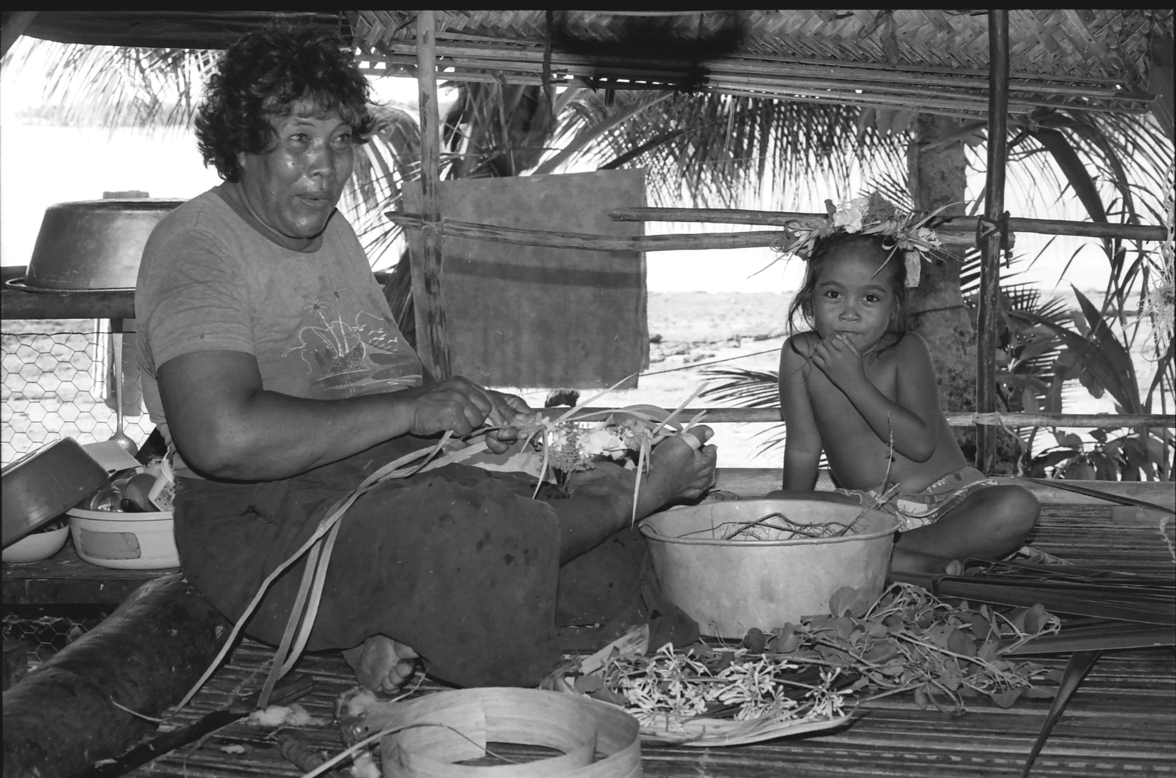 A woman and young daughter sitting inside a raised platform thatched building - making a woven mat, the daughter behind with her hand on her chin.