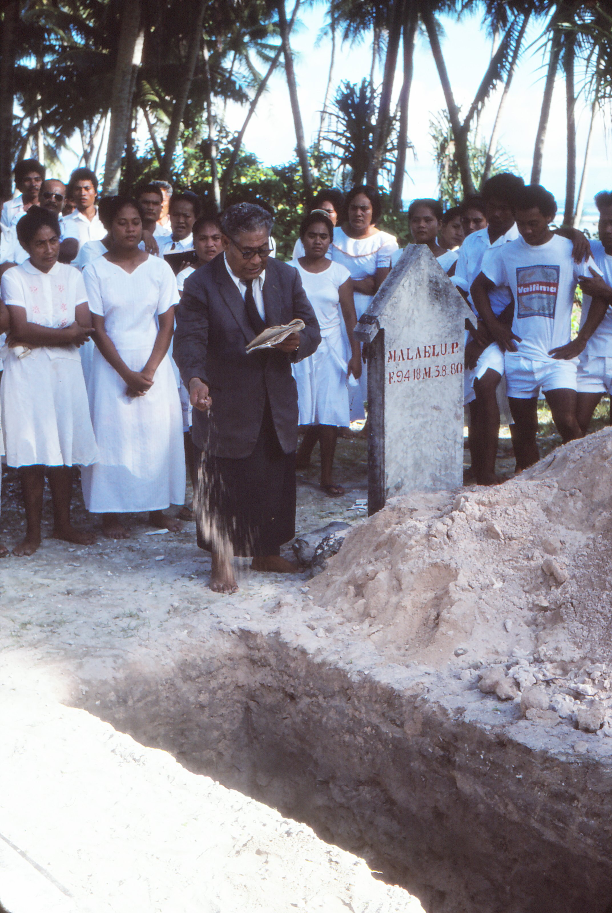A minister standing beside an open grave holding the bible open - performing a the funeral ceremony surrounded by people in white clothing, in a tropical outdoor setting with trees in the background.