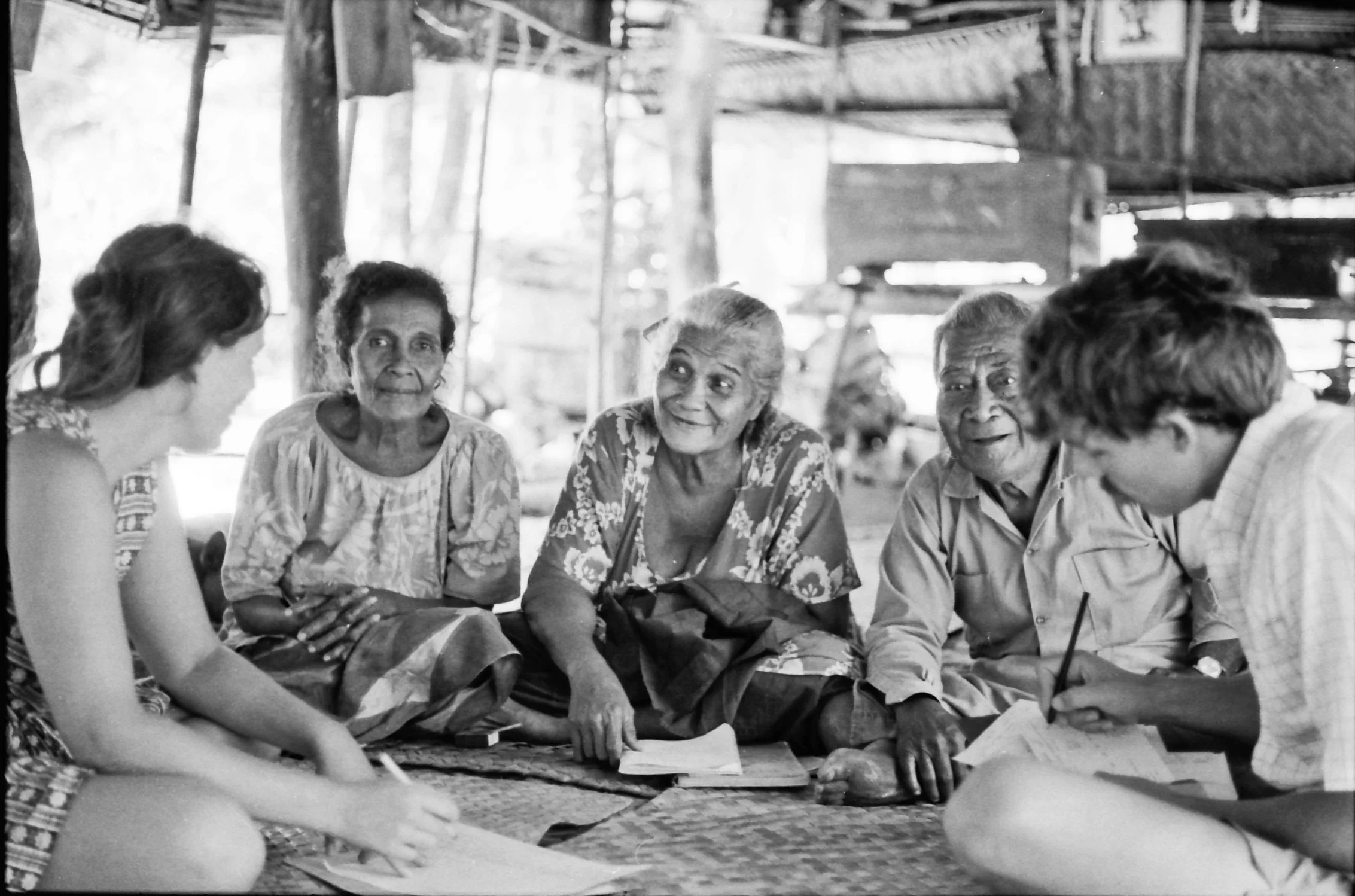 A group of five people, including elderly individuals and a younger woman, sitting on a mat and engaging in a conversation indoors, with some holding notebooks and pens.