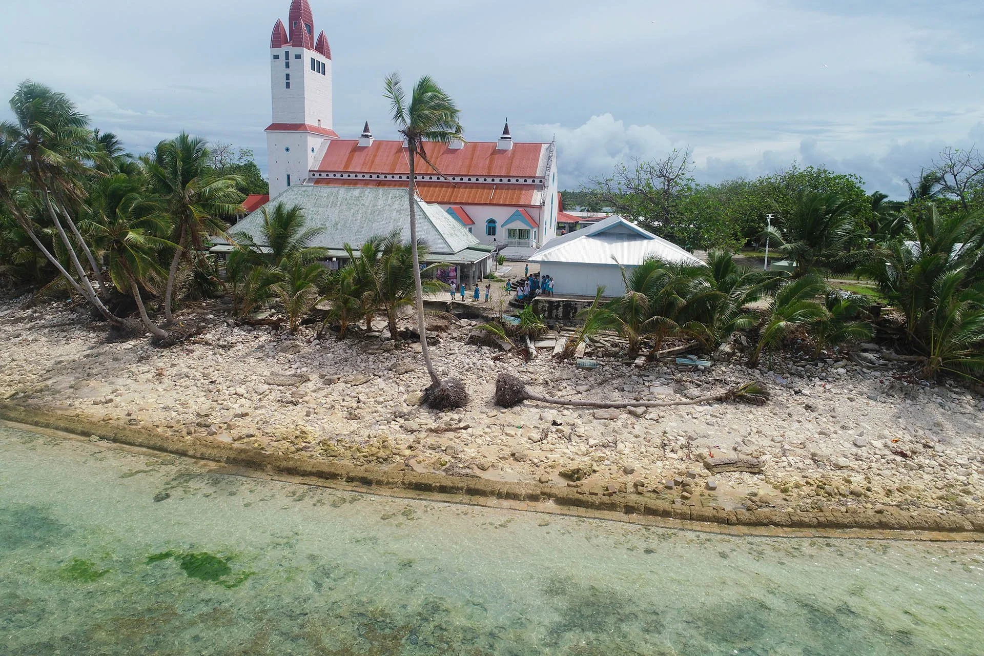 Nanumea's large white church with red roof taken from a drone just offshore, showing rocky and sandy shore, with cloudy skies overhead.