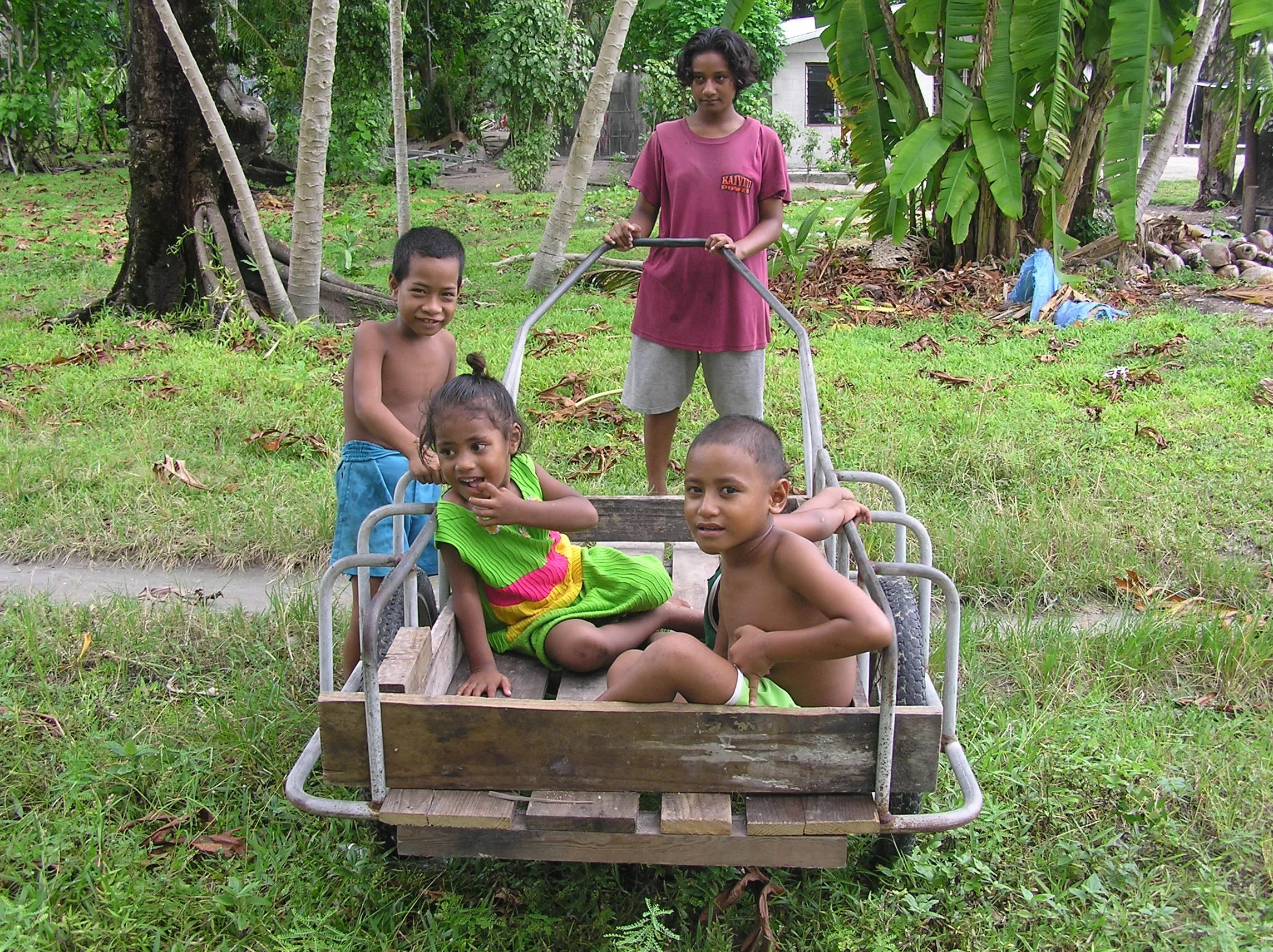 Four children, two boys and two girls, playing outdoors with a wooden cart in a lush green setting with trees and grass.