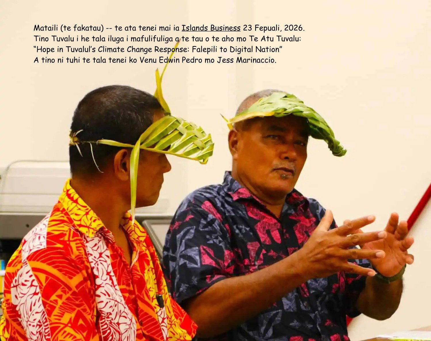 Two men wearing leafy hats and colorful shirts sitting indoors, with one man speaking and gesturing with his hands.