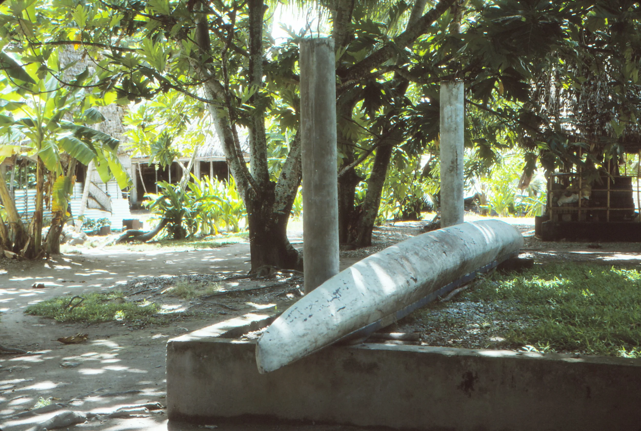 A large cement house foundation and an abandoned canoe hull turned upside down.  Lush, shaded area with trees and greenery..
