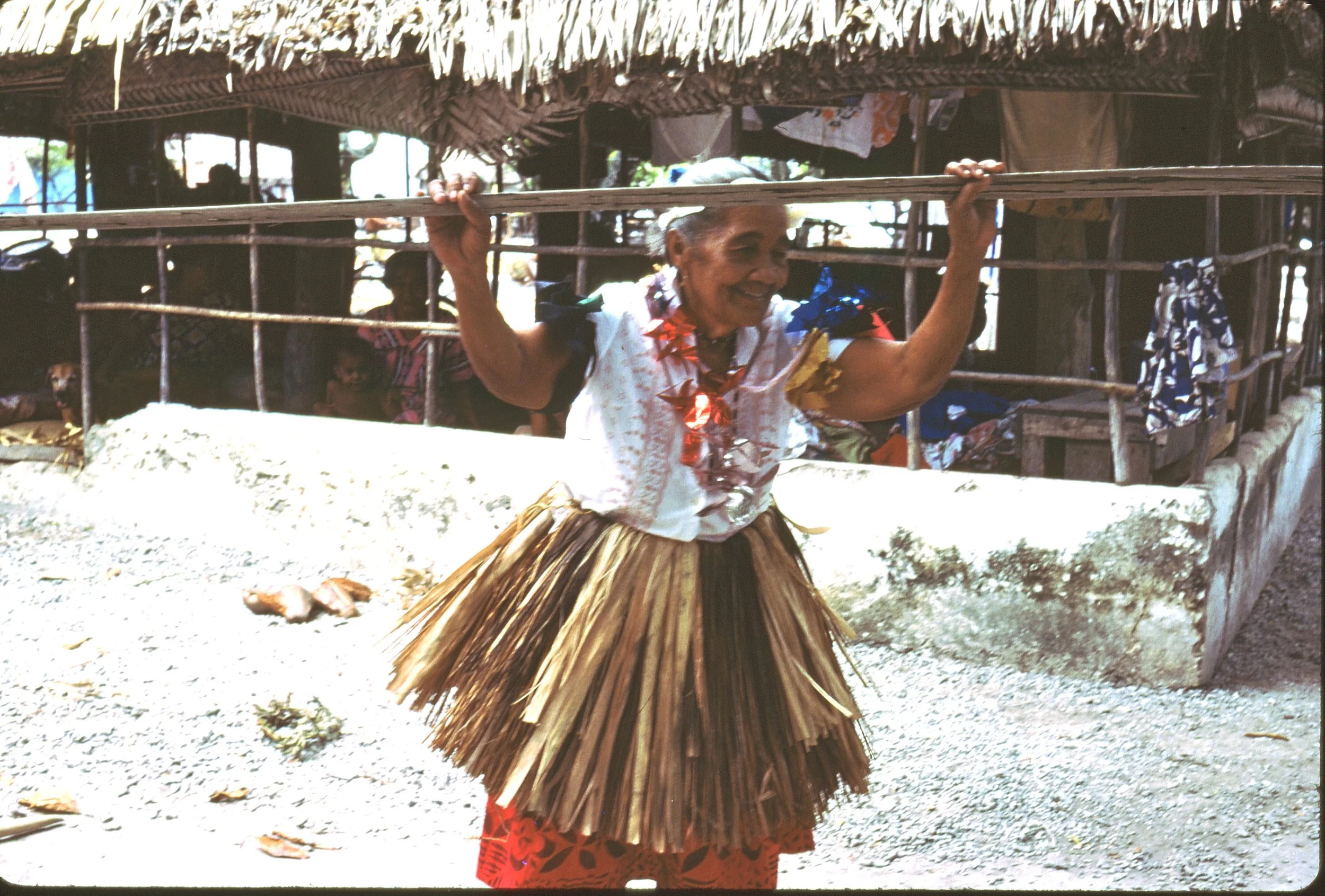 An elderly woman wearing a fiber dance skirt, a white blouse, and a lei, carrying a wooden staff  on her shoulders:  she is singing about the wedding of a grandchild.