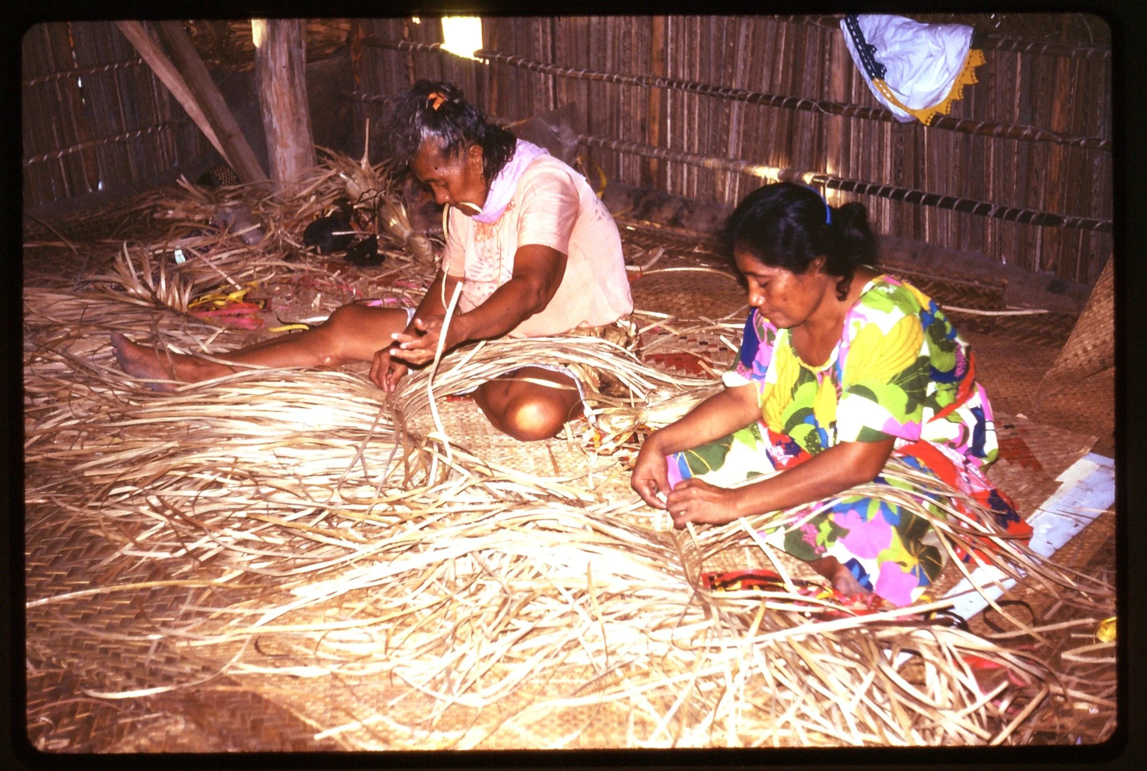 Two women weaving fine mats for the wedding to come of their grandchildren.  They are inside a traditional house.