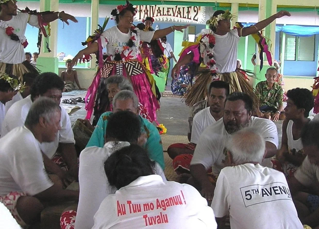 People participating in a traditional cultural dance and gathering indoors. Dancers are wearing grass skirts and floral accessories, performing on a raised platform, while the seated audience watches. The setting suggests a cultural event or celebration.