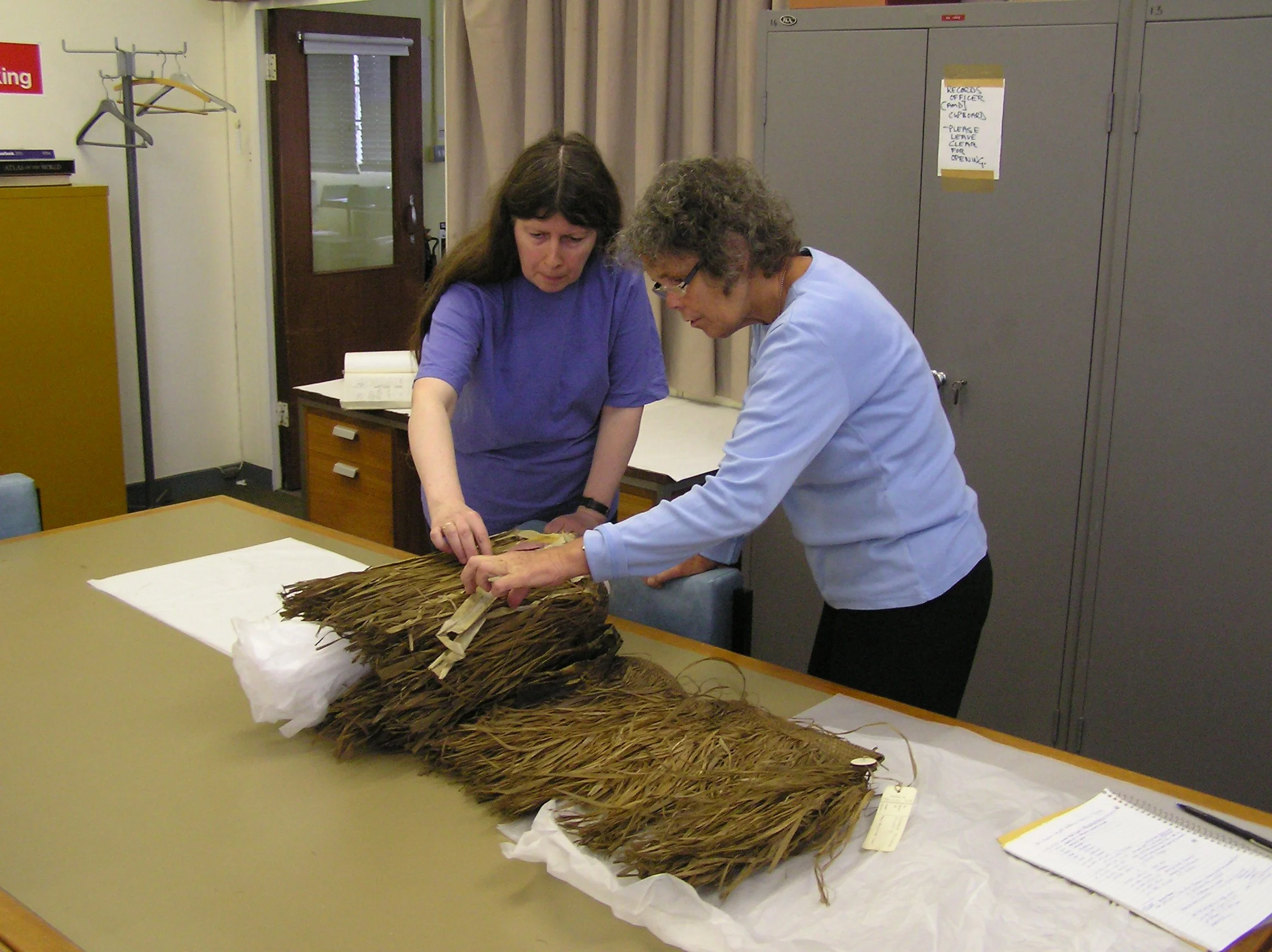 Two women examining a large bundle of dried plant material on a table, in a room with lockers and cabinets.