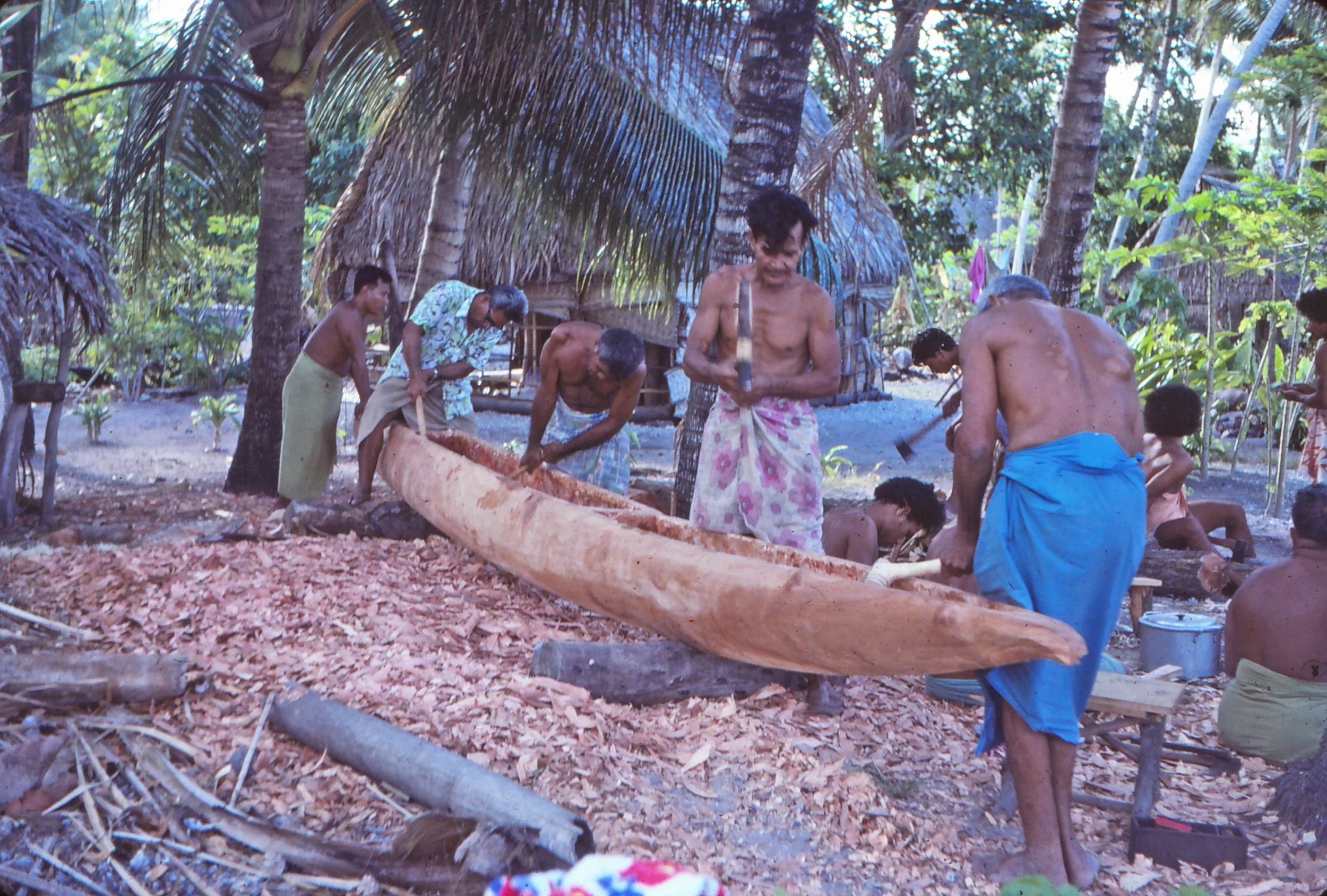 Group of men carving a large wooden canoe in a Nanumea's village shaded with palm trees.