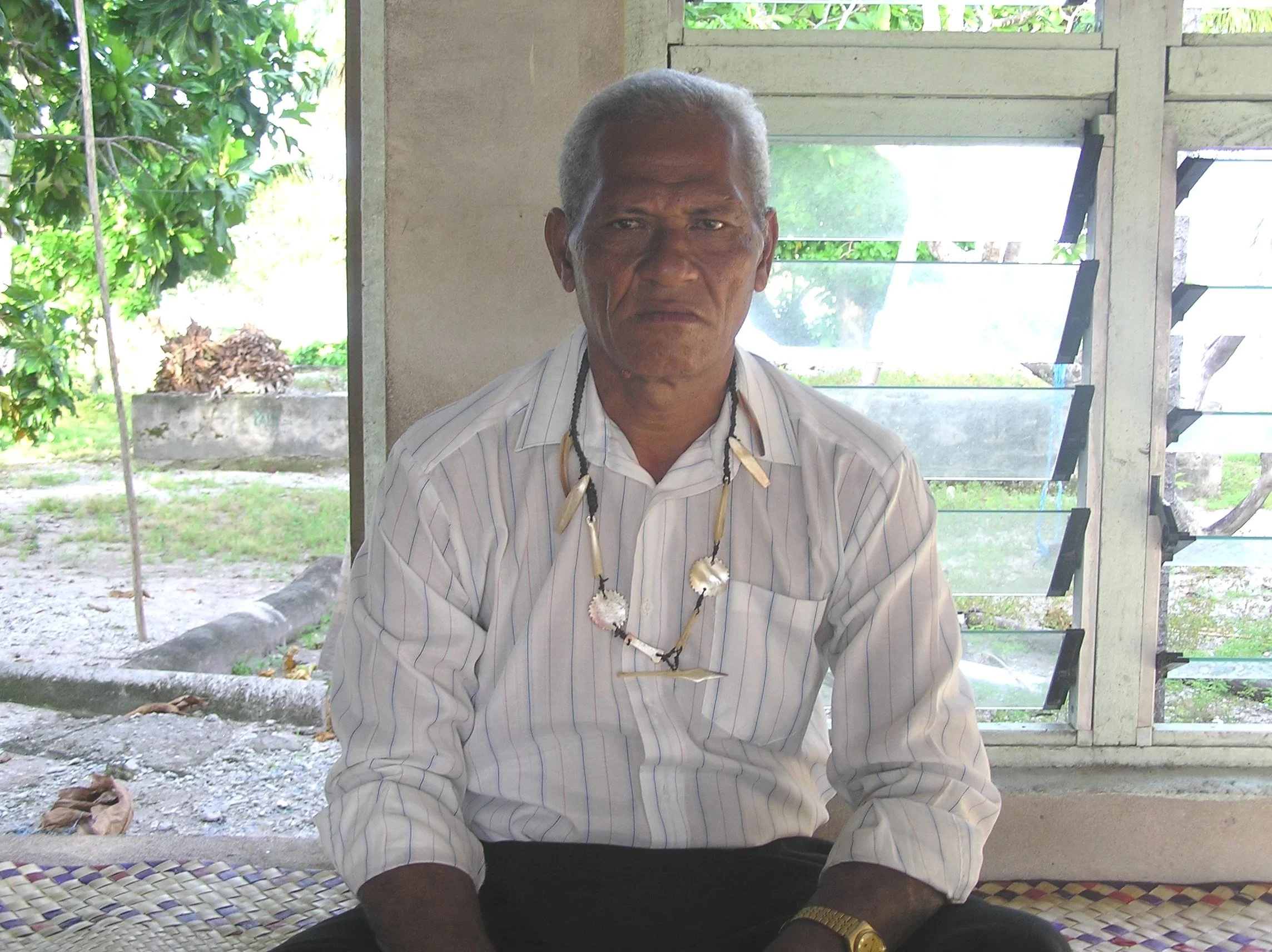 An elderly man with gray hair sitting indoors in front of a window with louvers, wearing a striped white shirt. He has a serious expression and is wearing a necklace with shells and a bracelet, with some greenery visible outside.