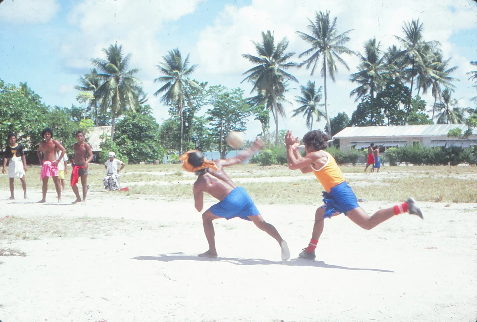 Pitching the Ano ball - two men in blue wrap around "hulu" -- on a sandy field with palm trees and houses in the background.