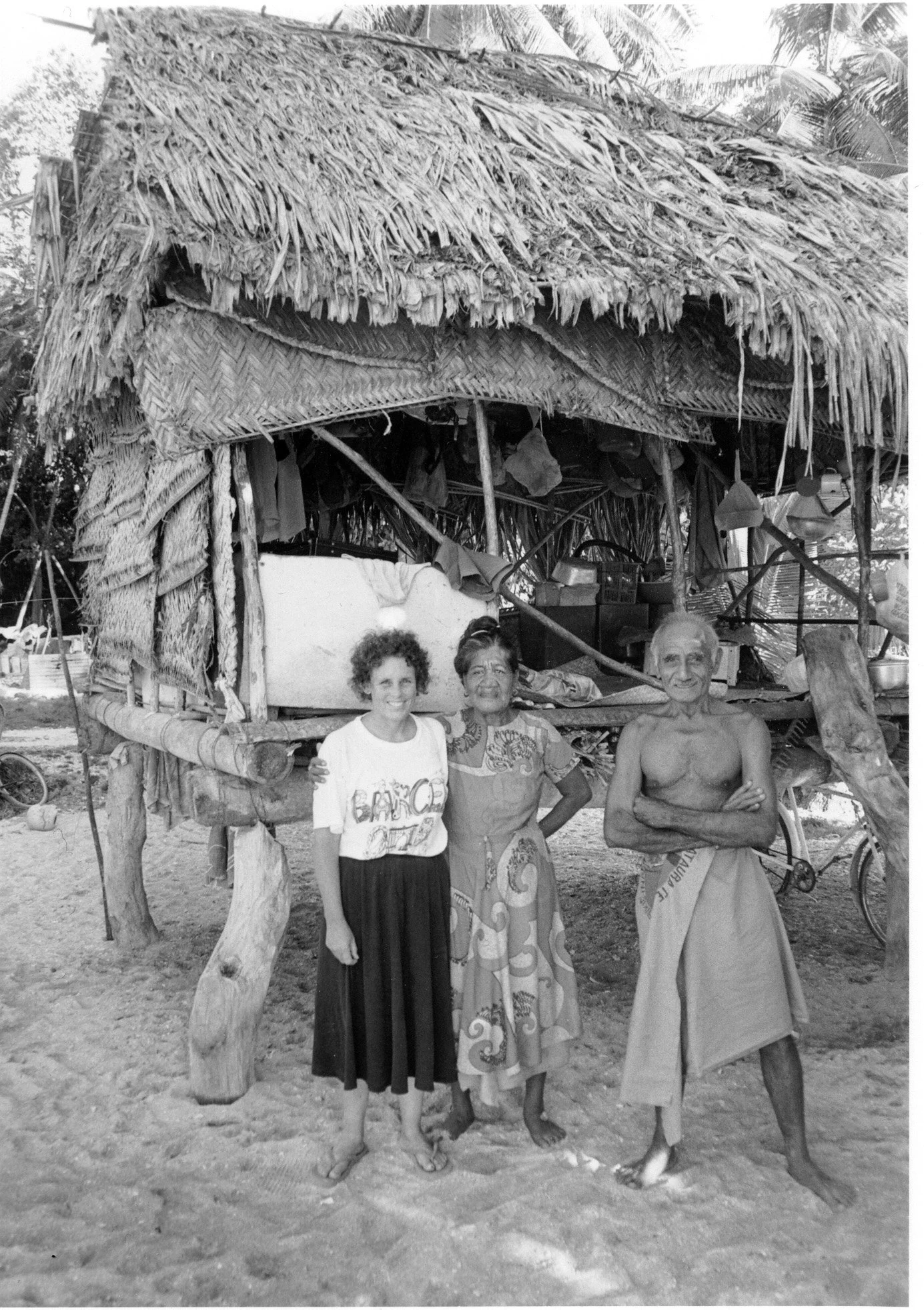 Anne Chambers with Salailoto and her husband at their modest thatched house, with trees and bicycles in the background.