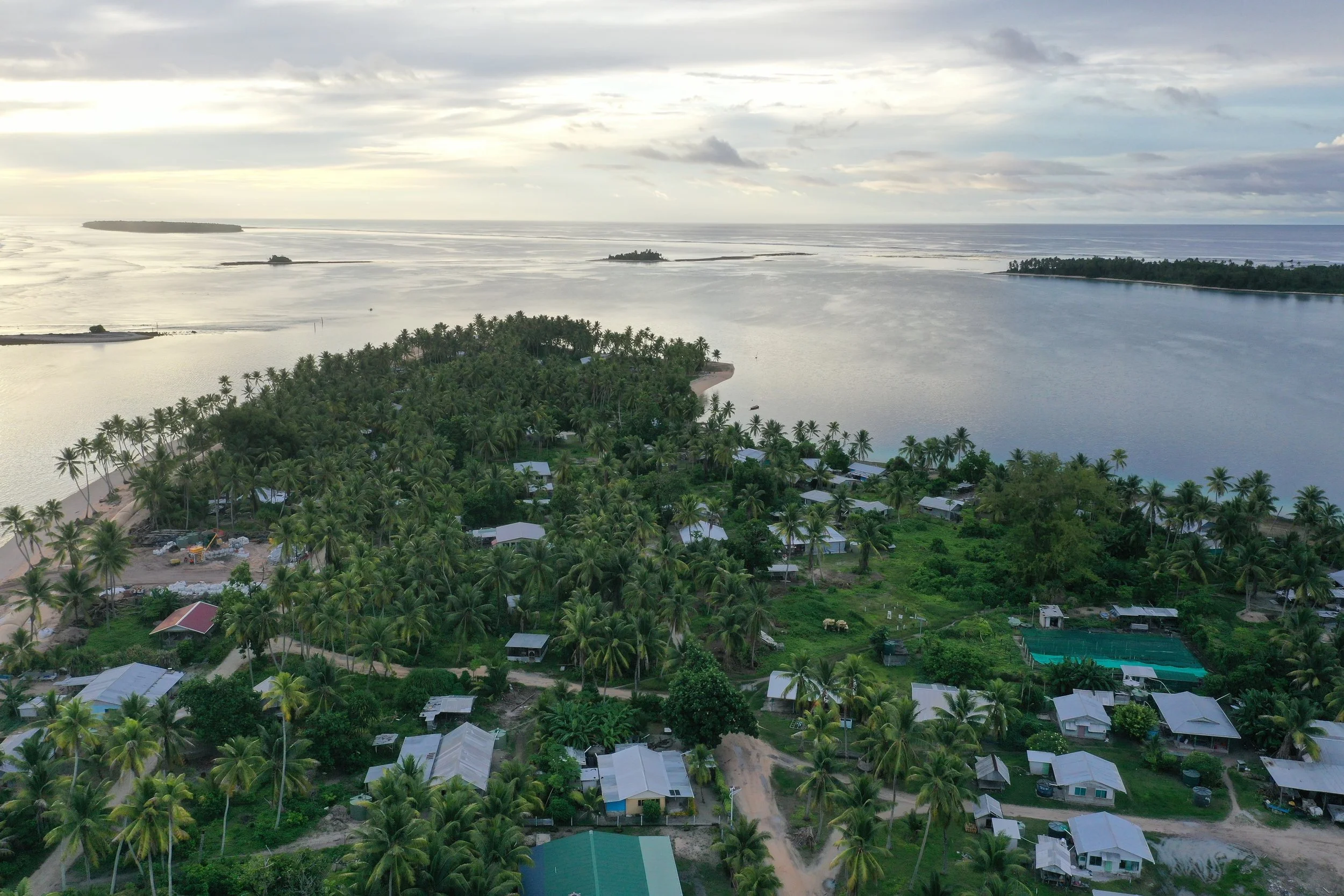 Aerial view to NW showing Hauma tip of Nanumea village, with numerous palm trees, scattered houses, and sandy beaches, surrounded by calm water and distant small islands under a cloudy sky.