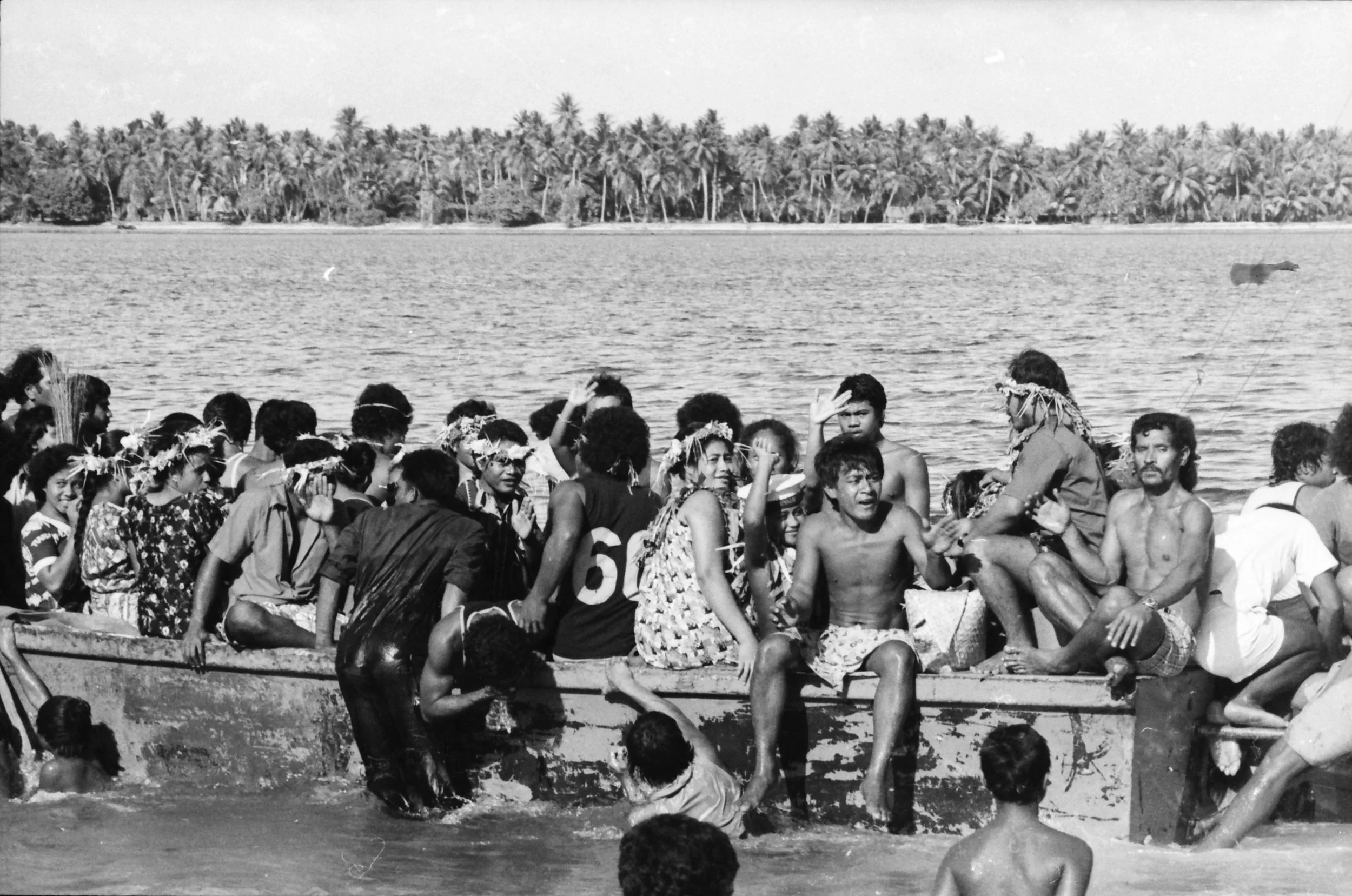 A black and white photo of a large group of people leaving Nanumea's lagoon on a boat, some wearing floral headbands, with some standing in the water beside the boat.  Lagoon and palm trees in the background.