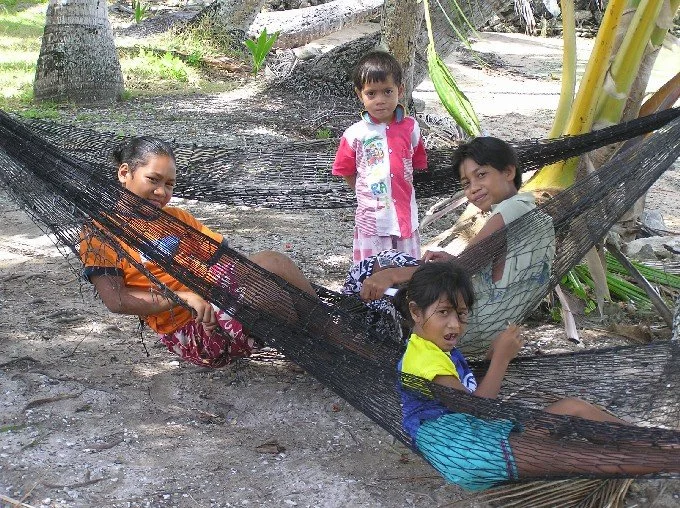 Four children are playing on a hammock tied between trees on a sandy ground.