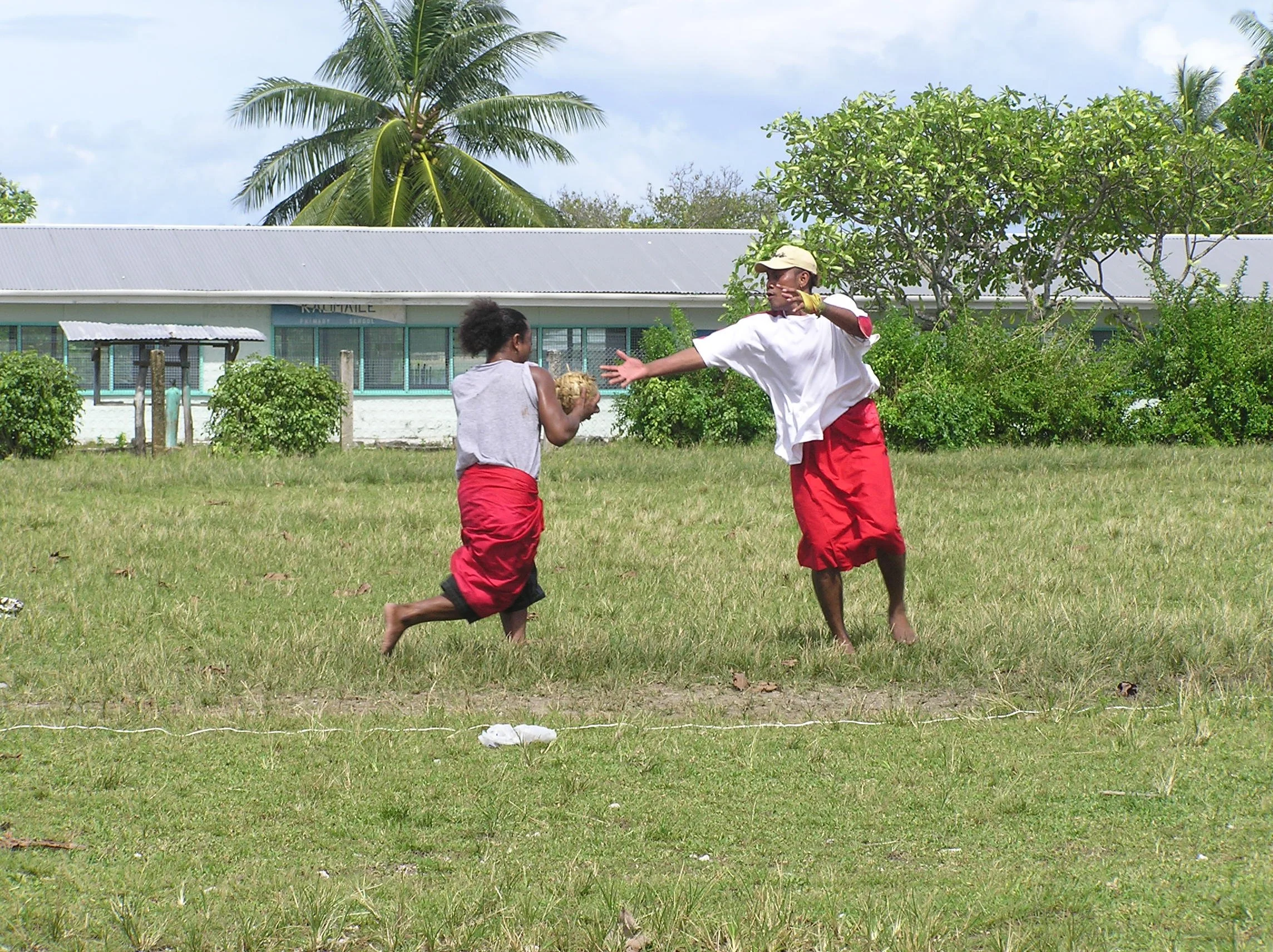 Young men hurling the Ano ball on a grassy field, one reaching to connect and hurl the ball, both dressed in red hulu wrap arounds and tee shirts, with trees and a school building in the background.