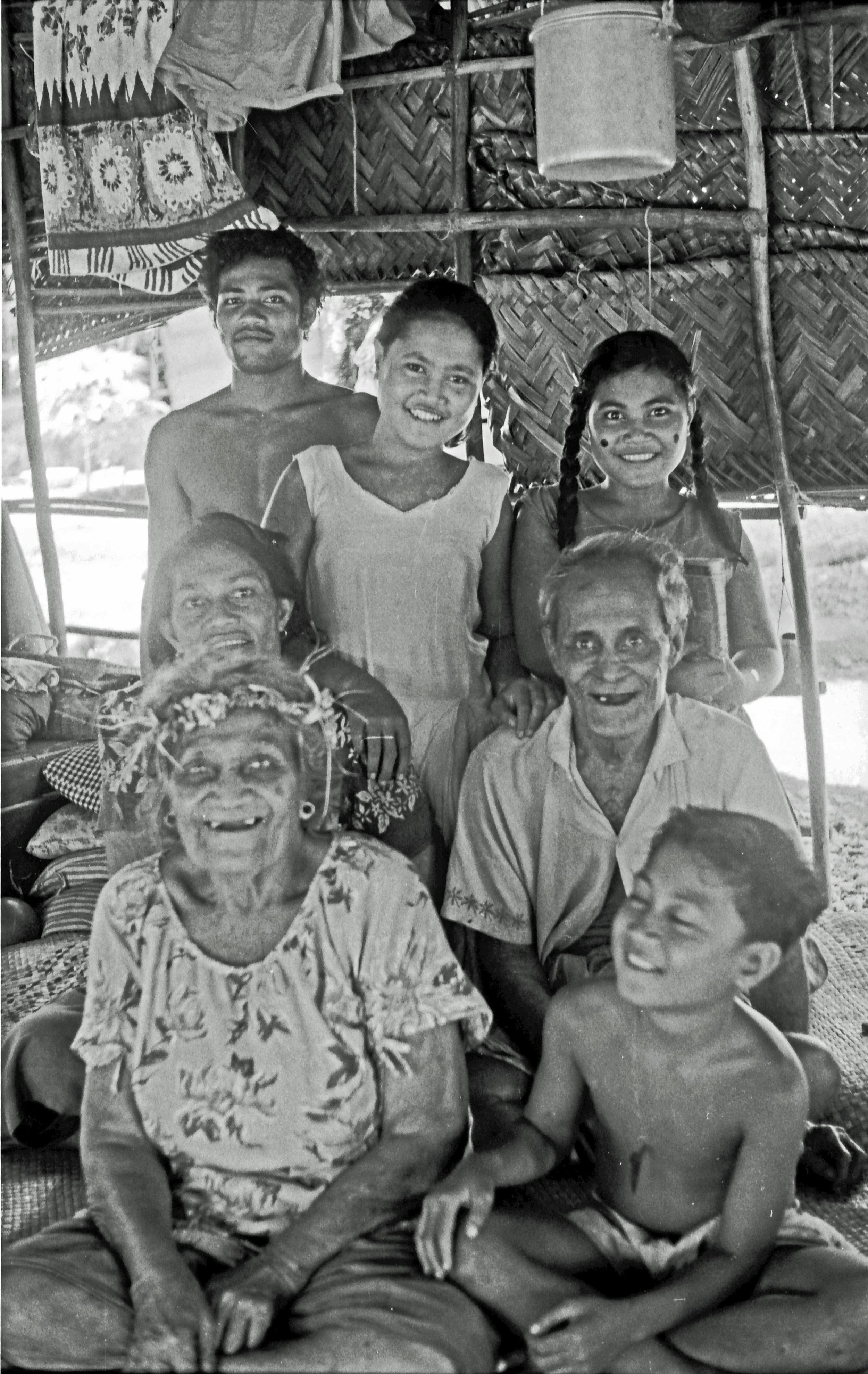 A black-and-white photo of a multigenerational family gathered inside a village house, with woven walls and some fabric hanging overhead. The group are all smiling and posing for the camera.