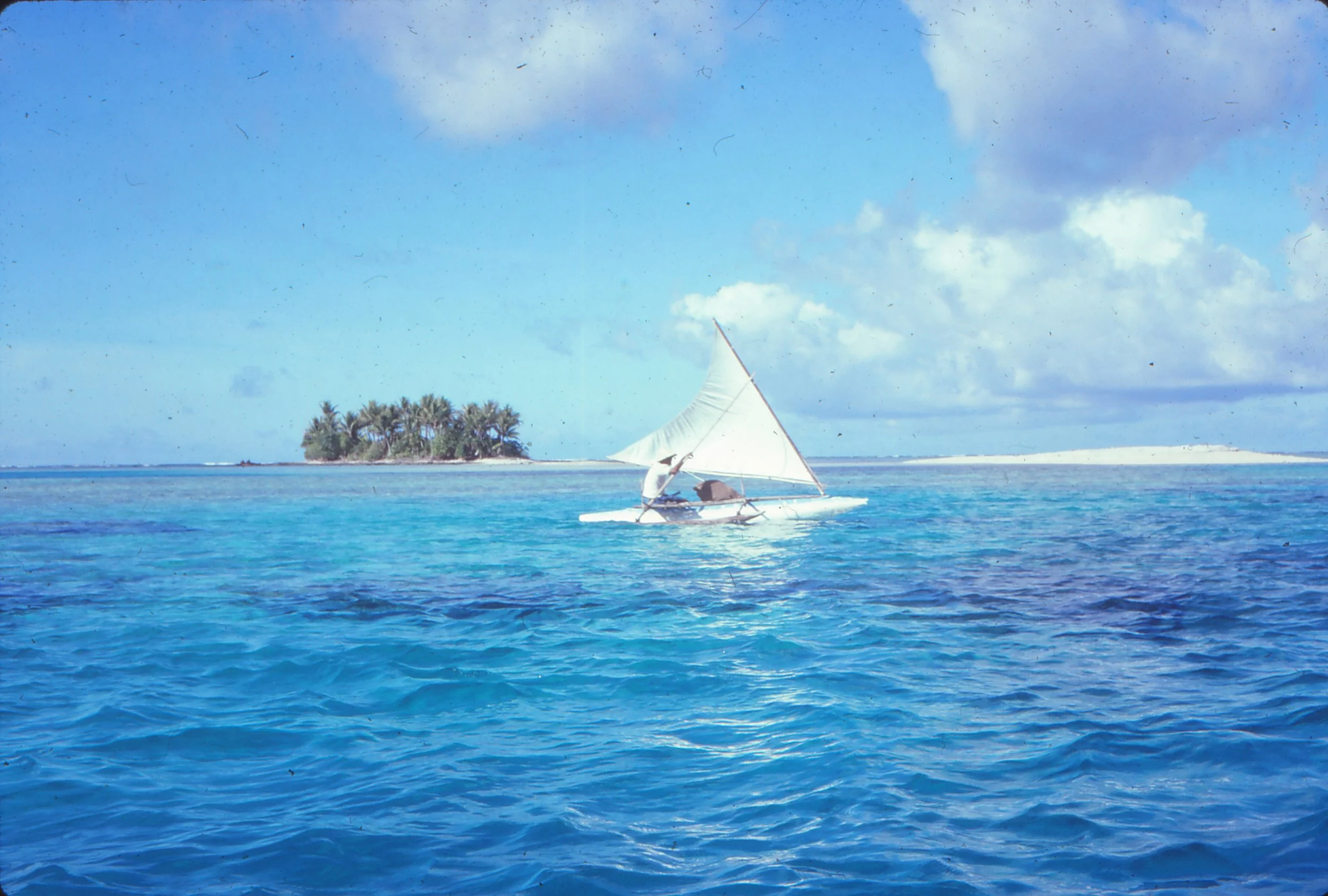 A small single person canoe with a white sail goes through Haufaga pass on its way to Lakena.  Blue ocean water and in the rear a small island with palm trees, under a partly cloudy sky.