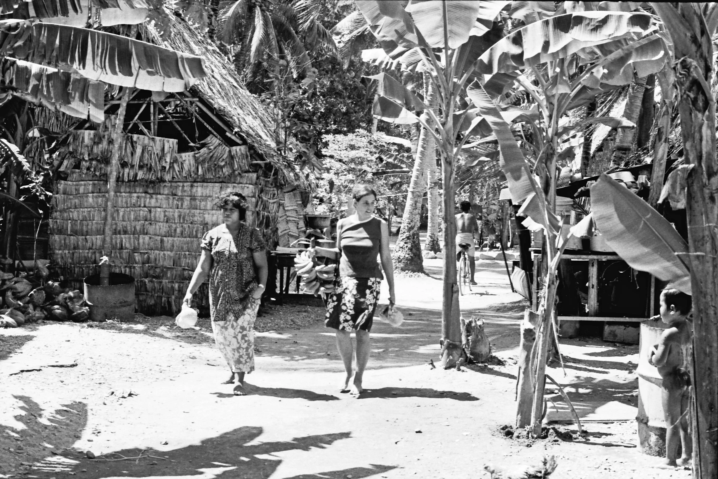 Anne and Sunema walking walk through Nanumea village with backdrop of banana trees, thatched houses, and villagers engaged in daily activities.