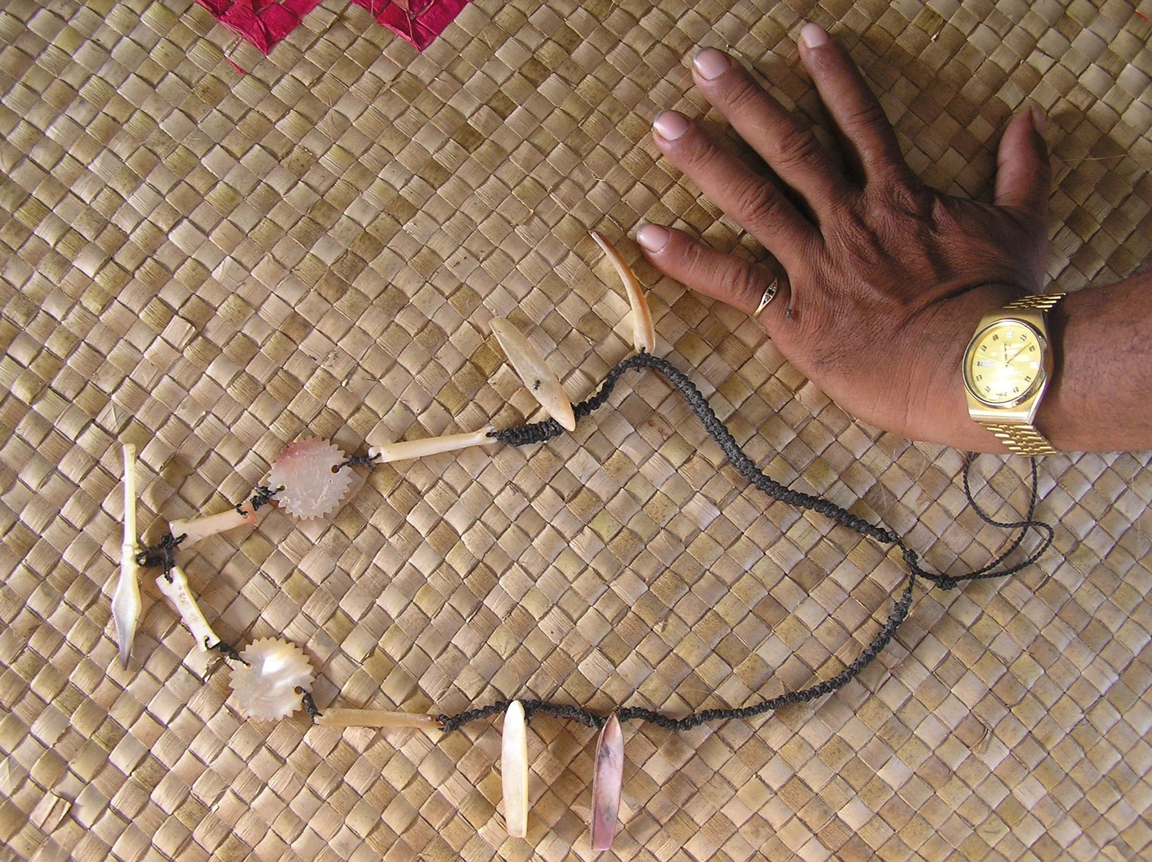 A person's hand rests on a woven surface, next to a necklace made of small black beads with various shell and bone pendants.