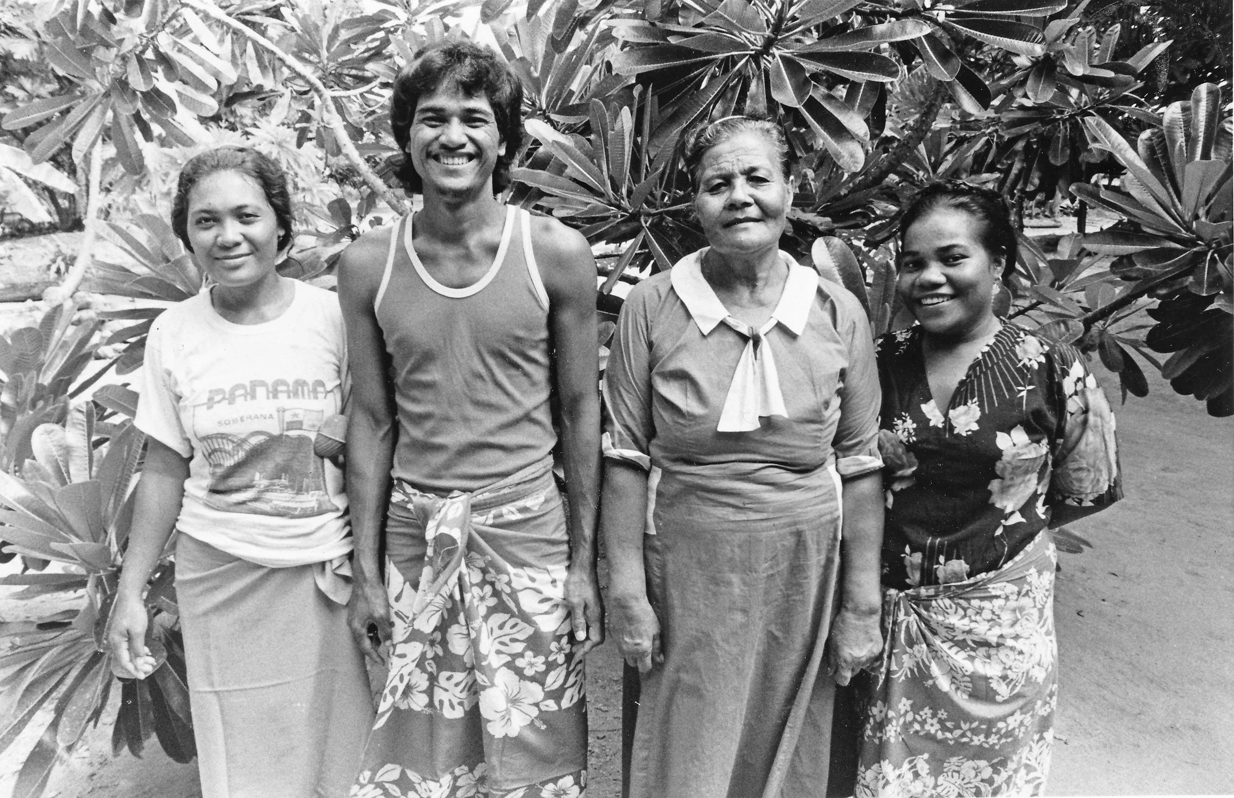 Black and white photo of four people standing outdoors in front of large leafy plants. The group includes two women and two younger women, all smiling and dressed in casual, traditional, and floral clothing.