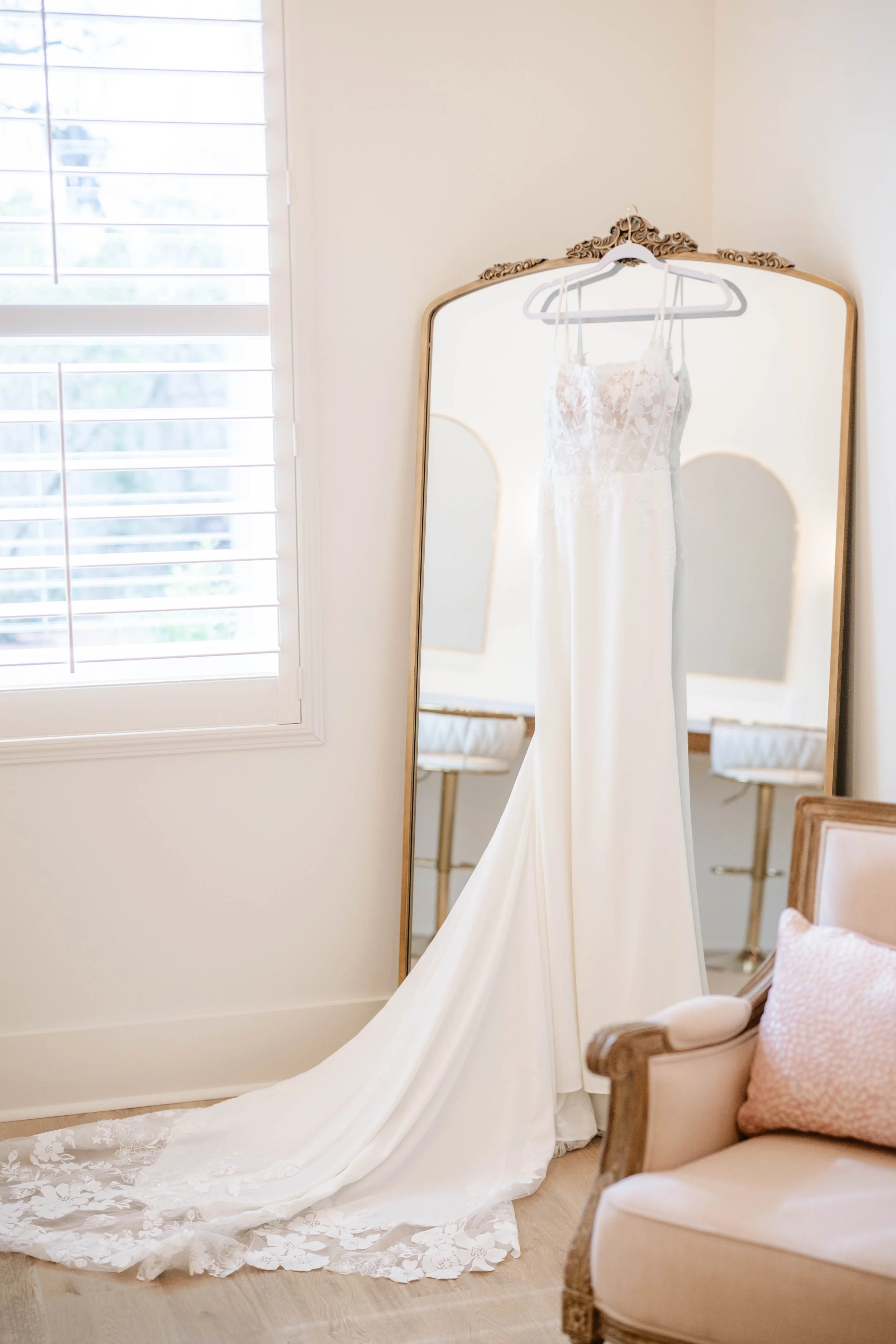 Elegant white wedding dress with lace train on a hanger, reflected in a large ornate mirror in Willow Point Venue's softly decorated bridal suite.