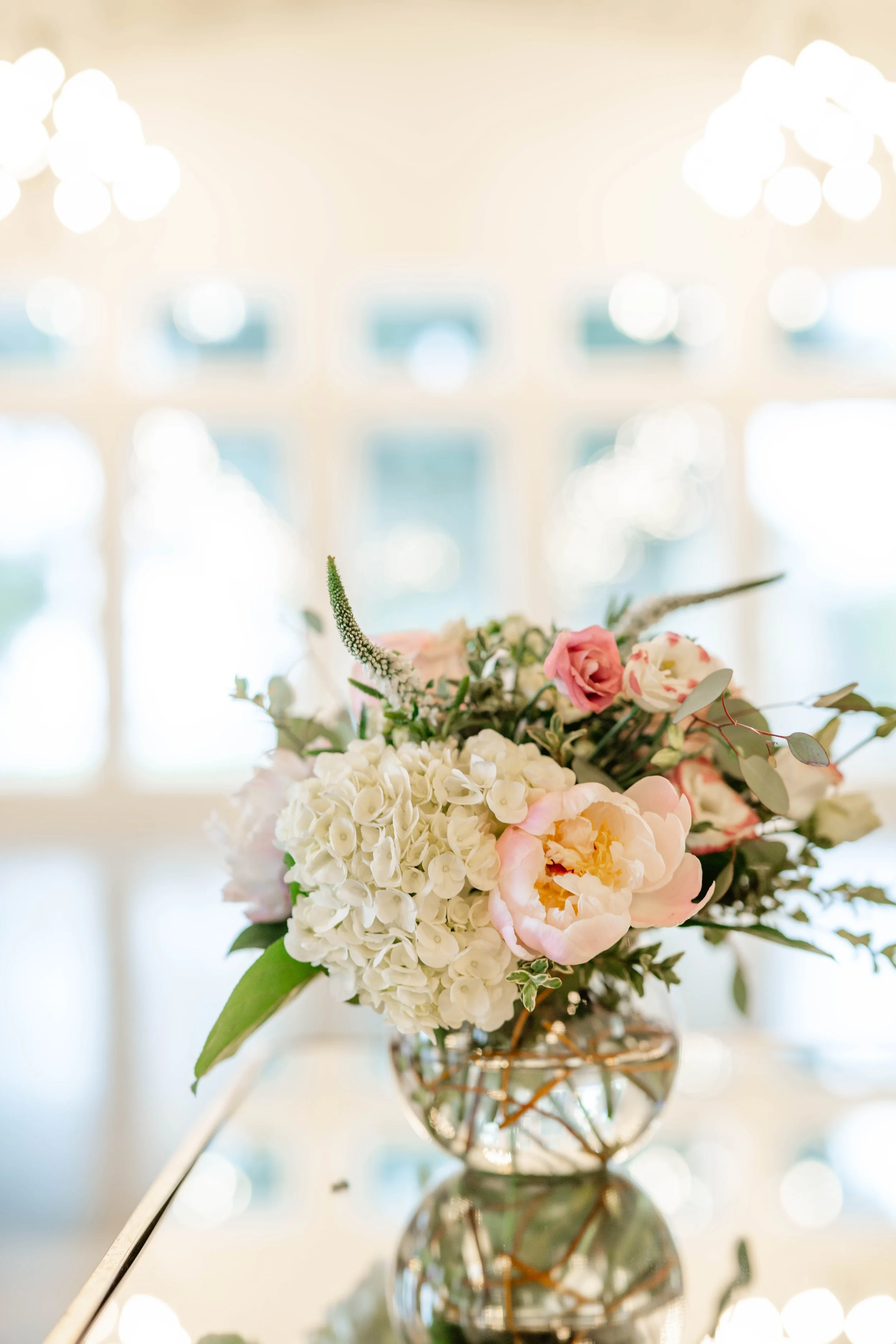Elegant floral arrangement with white hydrangeas, pink peonies, and greenery in a glass vase on a reflective surface.