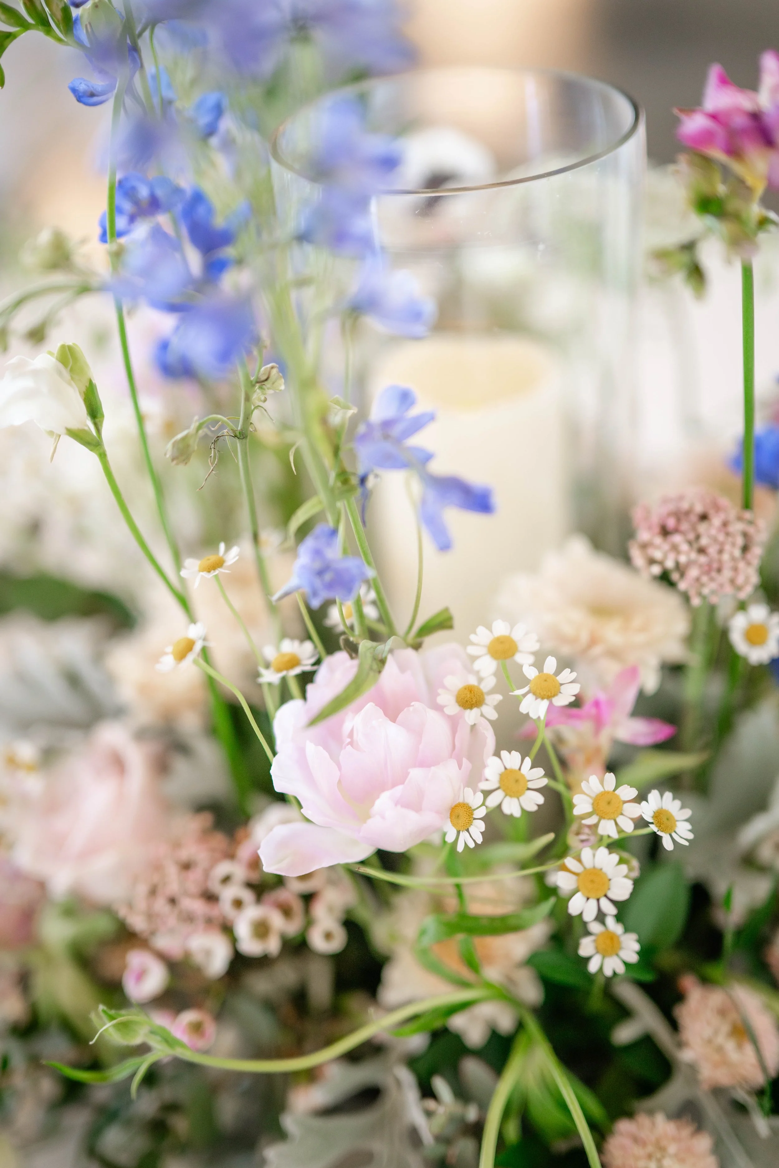 Close-up of a floral arrangement with pink peonies, blue delphiniums, white daisies, and green foliage surrounding a glass candle holder.