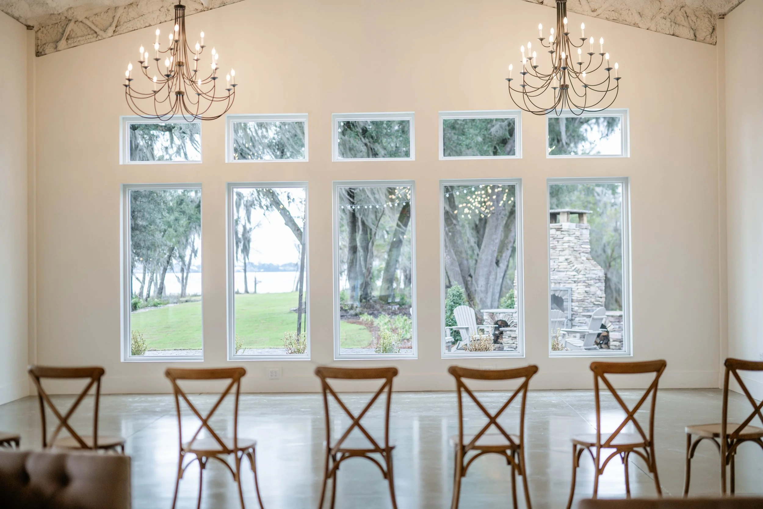 Interior of Willow Point Venue's spacious room with large windows overlooking a garden, brown chairs aligned in a row, and two chandeliers hanging from the ceiling.