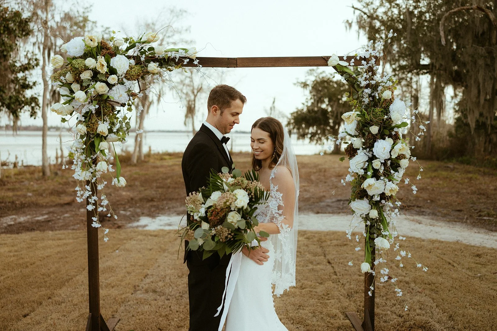Bride and groom standing under a wooden arch decorated with white flowers, greenery around, outdoor setting at Willow Point Venue.