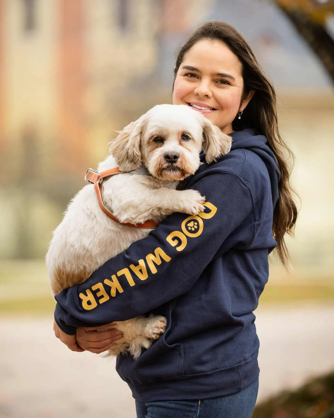 A woman smiling while holding a small dog in her arms outdoors with blurred fall foliage in the background.