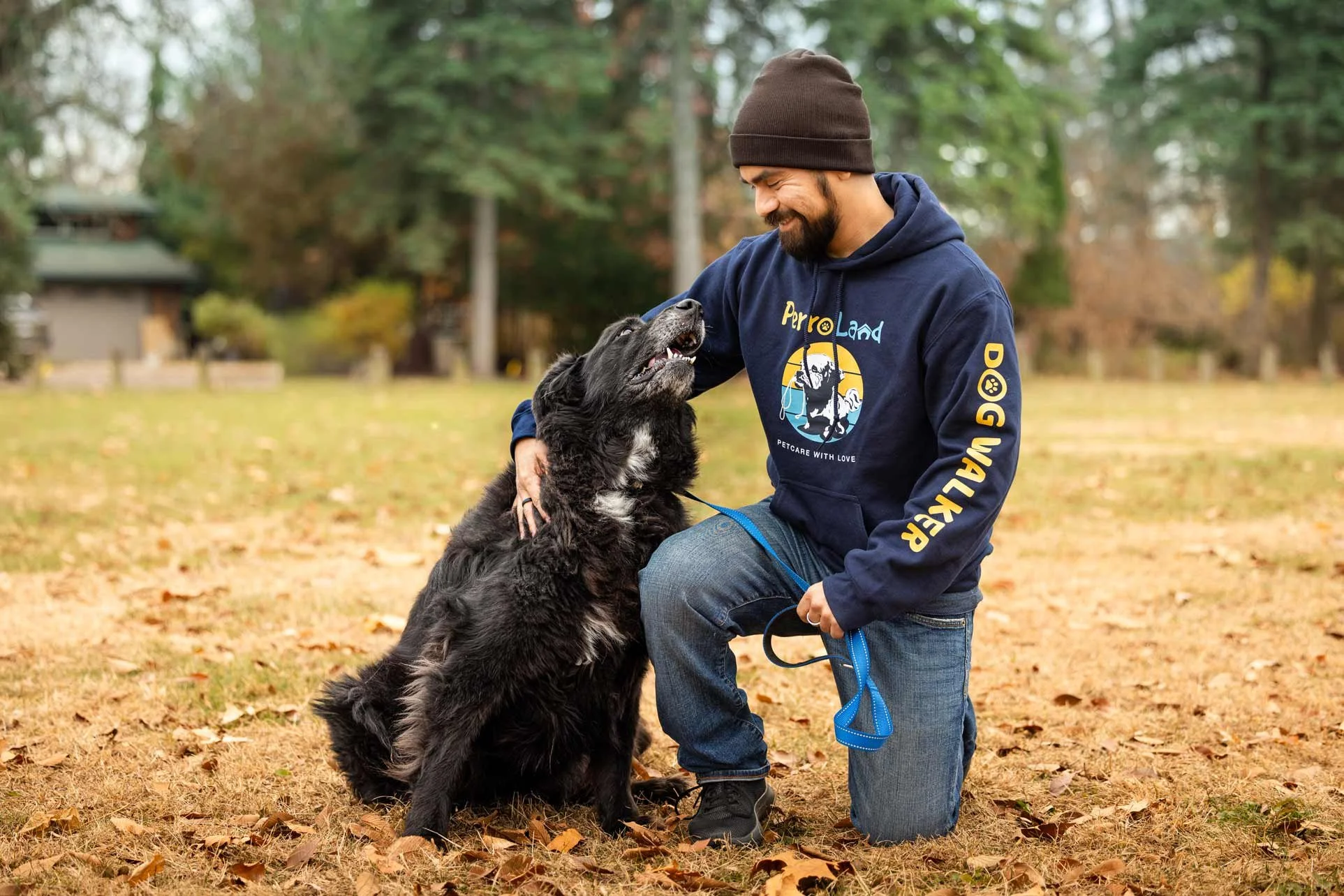A man kneeling in a park with fallen leaves, smiling and playing gently with a large black and white dog, both showing affection.