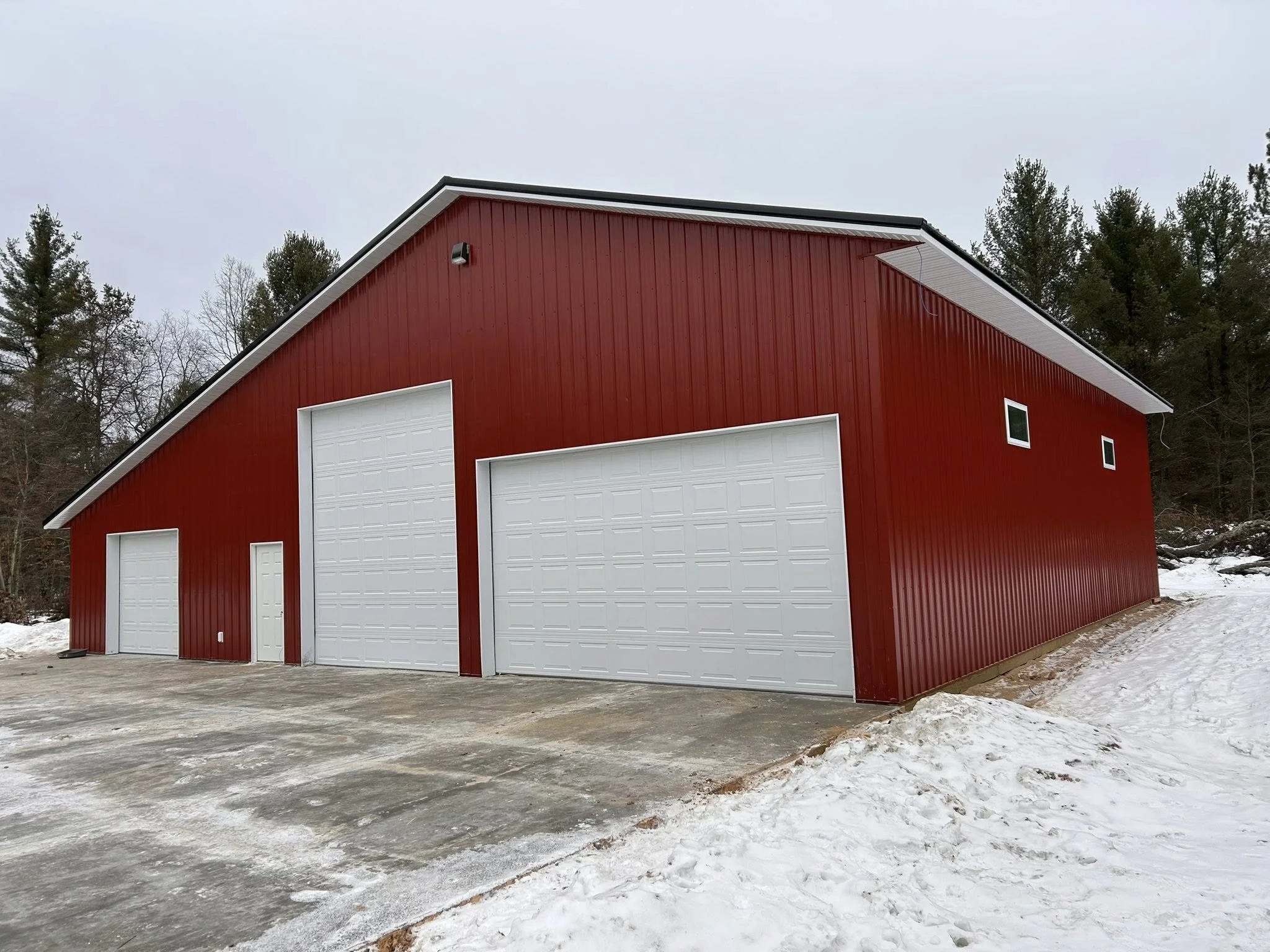 Red metal barn with three white garage doors and a small side door, situated on a snowy ground with trees in the background.