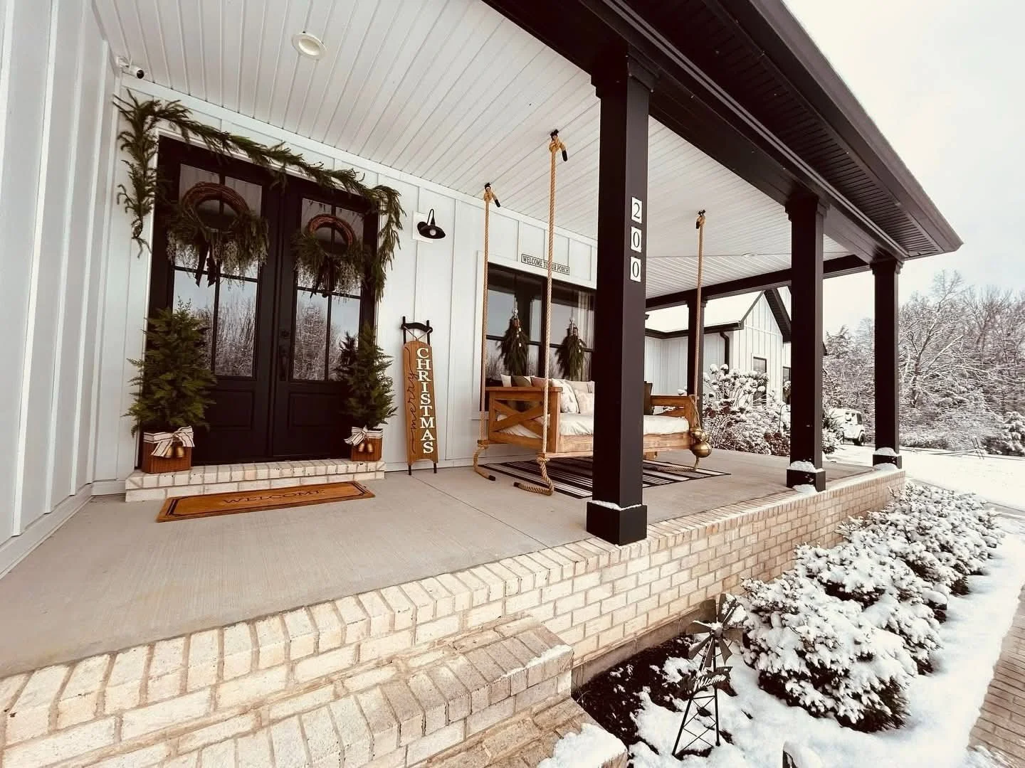 Christmas decorated porch with black doors, greenery, and a swing in a snowy yard.