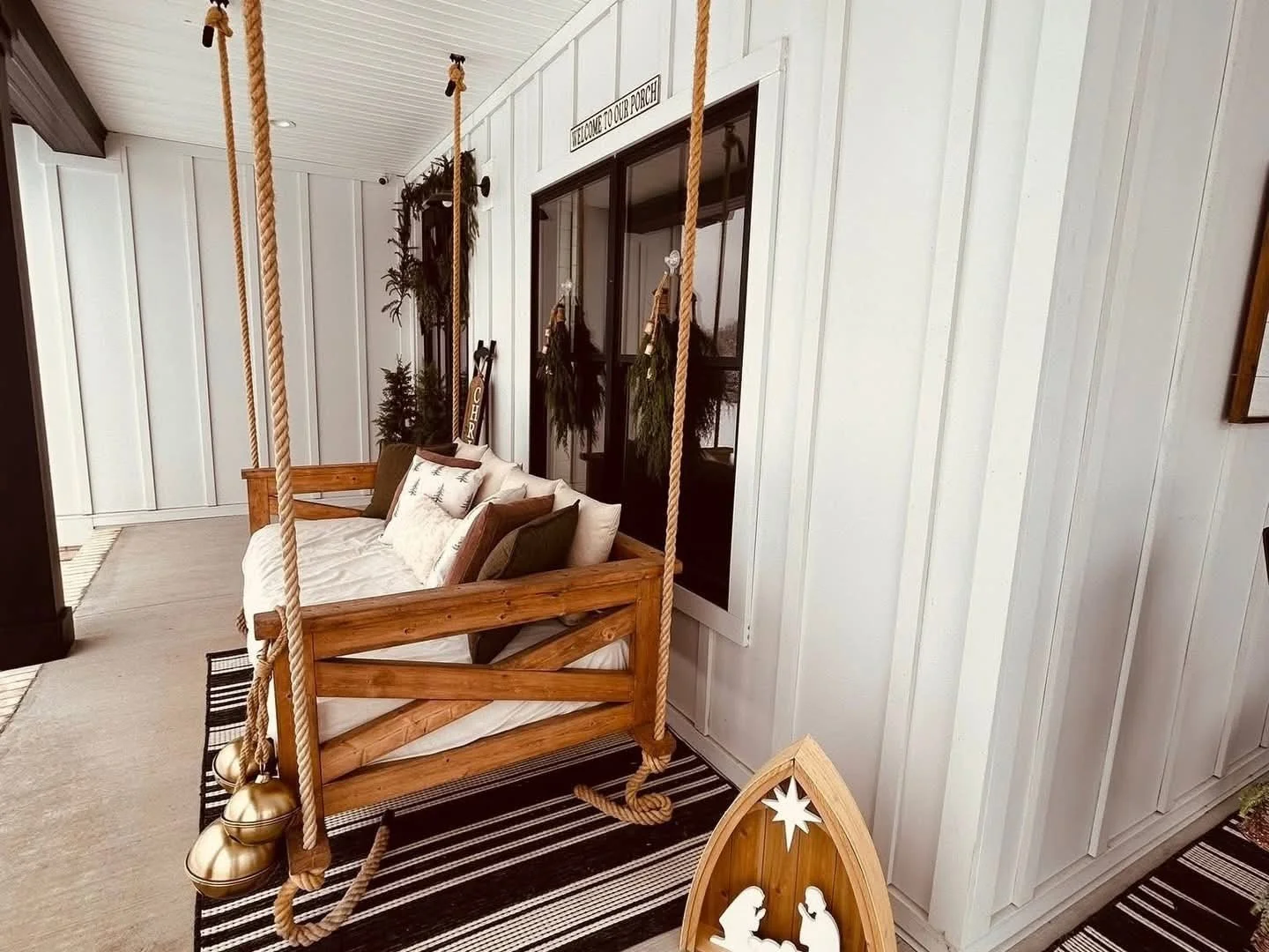 A porch decorated with Christmas decor, featuring a wooden swing with cushions, hanging ropes, holiday wreaths, and a nativity scene ornament on a rug.
