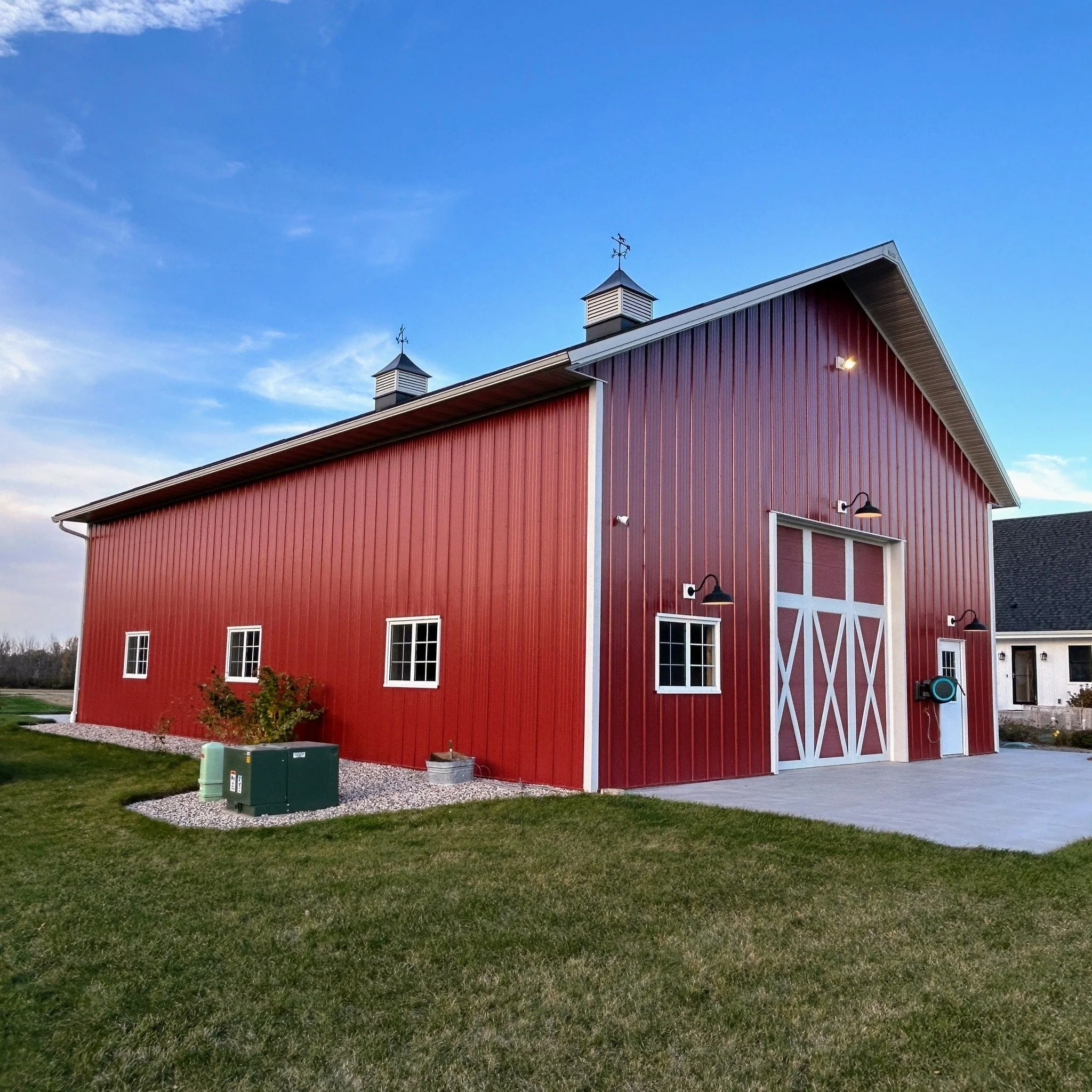 Red barn with white trim, small windows, and double doors with crossbuck design, situated on a grassy lawn with a concrete driveway, under a clear blue sky.
