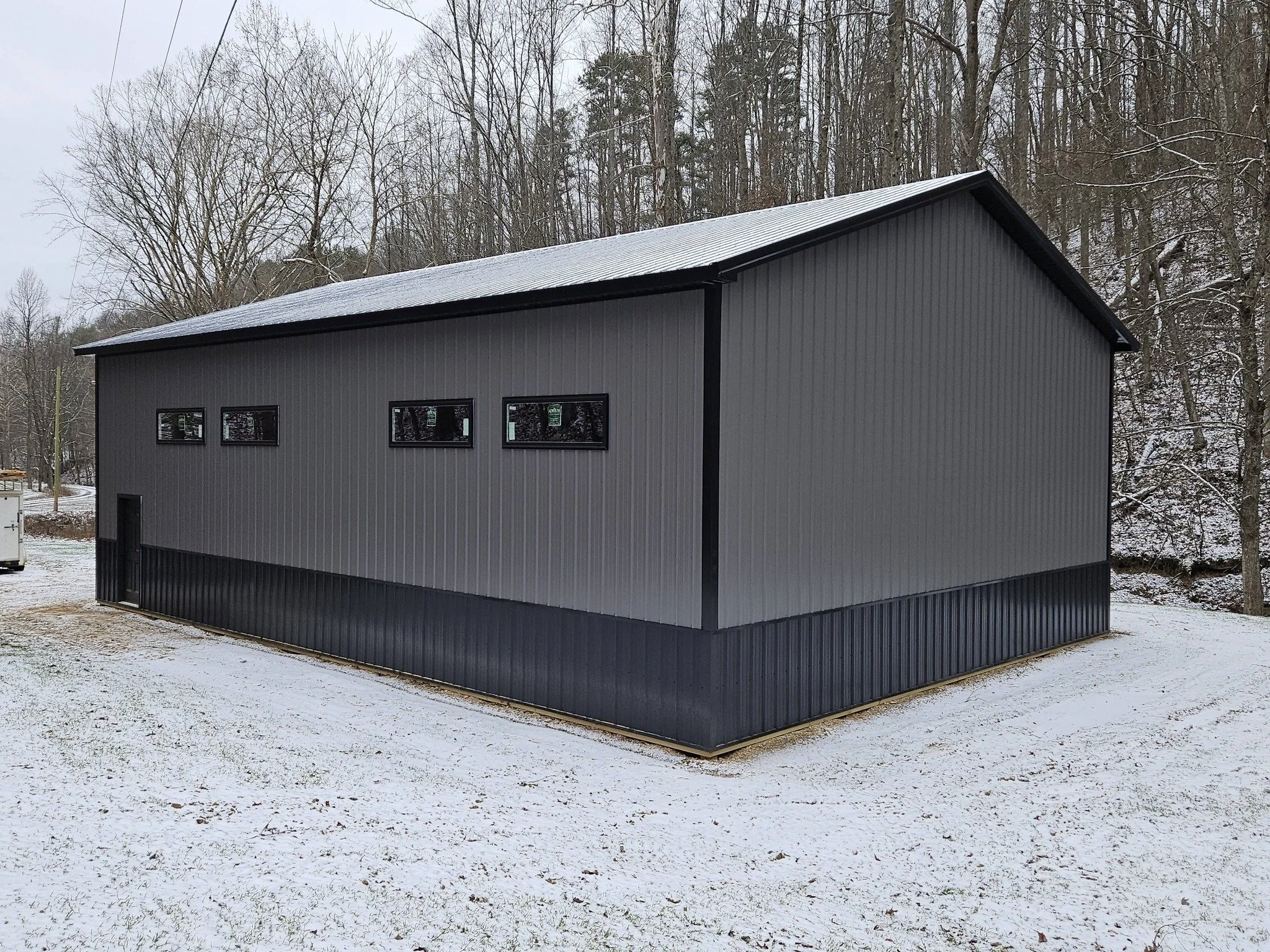 A standalone gray metal building with a sloped roof and small rectangular windows, situated on a snow-covered ground with leafless trees in the background.