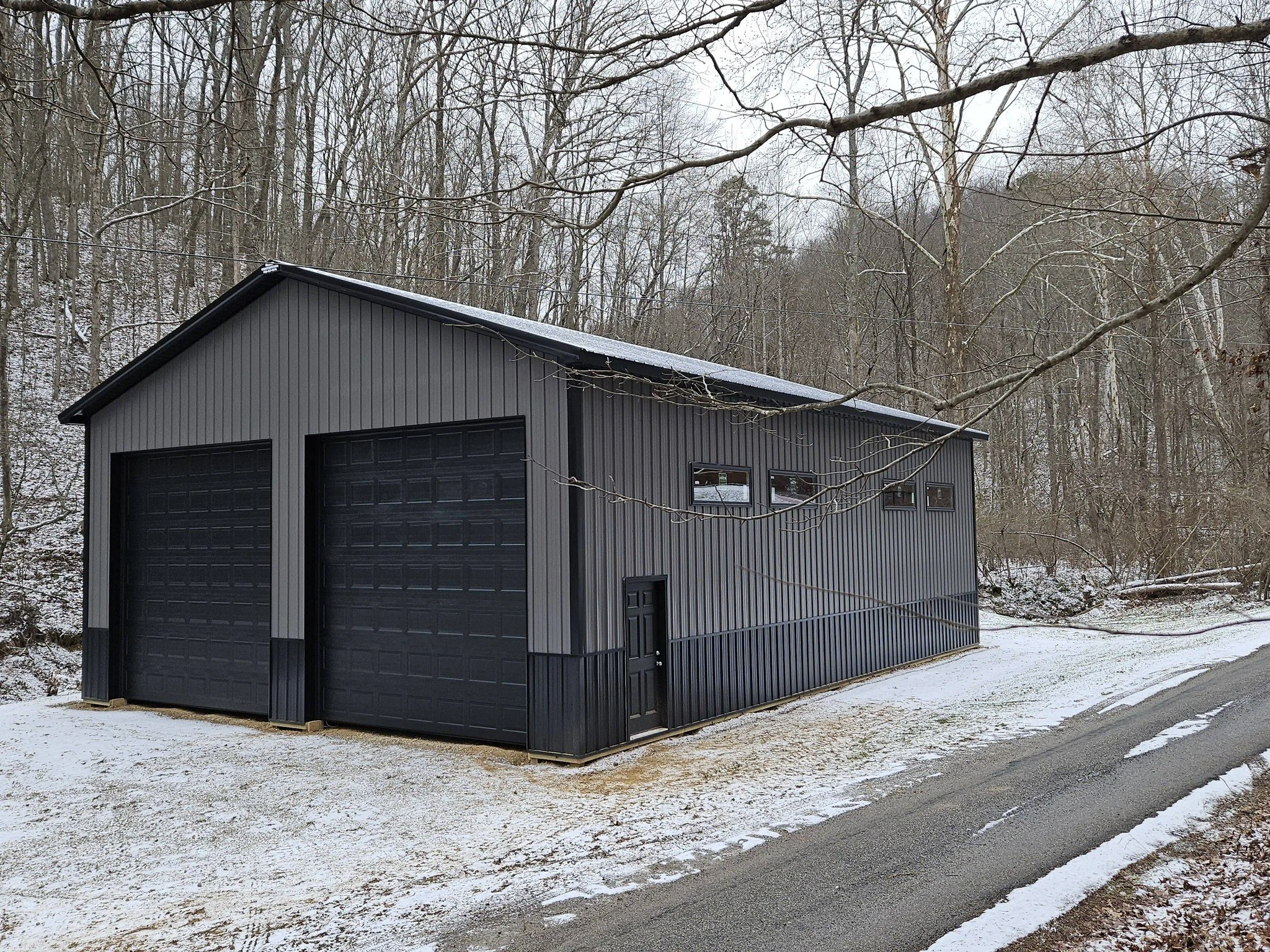 A black metal garage with two large garage doors and a small pedestrian door, situated in a snowy wooded area.
