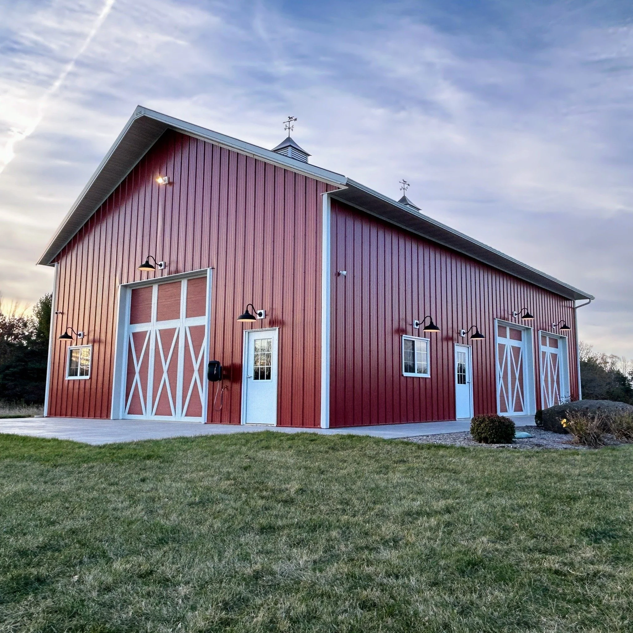 Red barn with white trim and large sliding doors, surrounded by a grassy lawn and small shrubs, under a cloudy sky in the early evening.