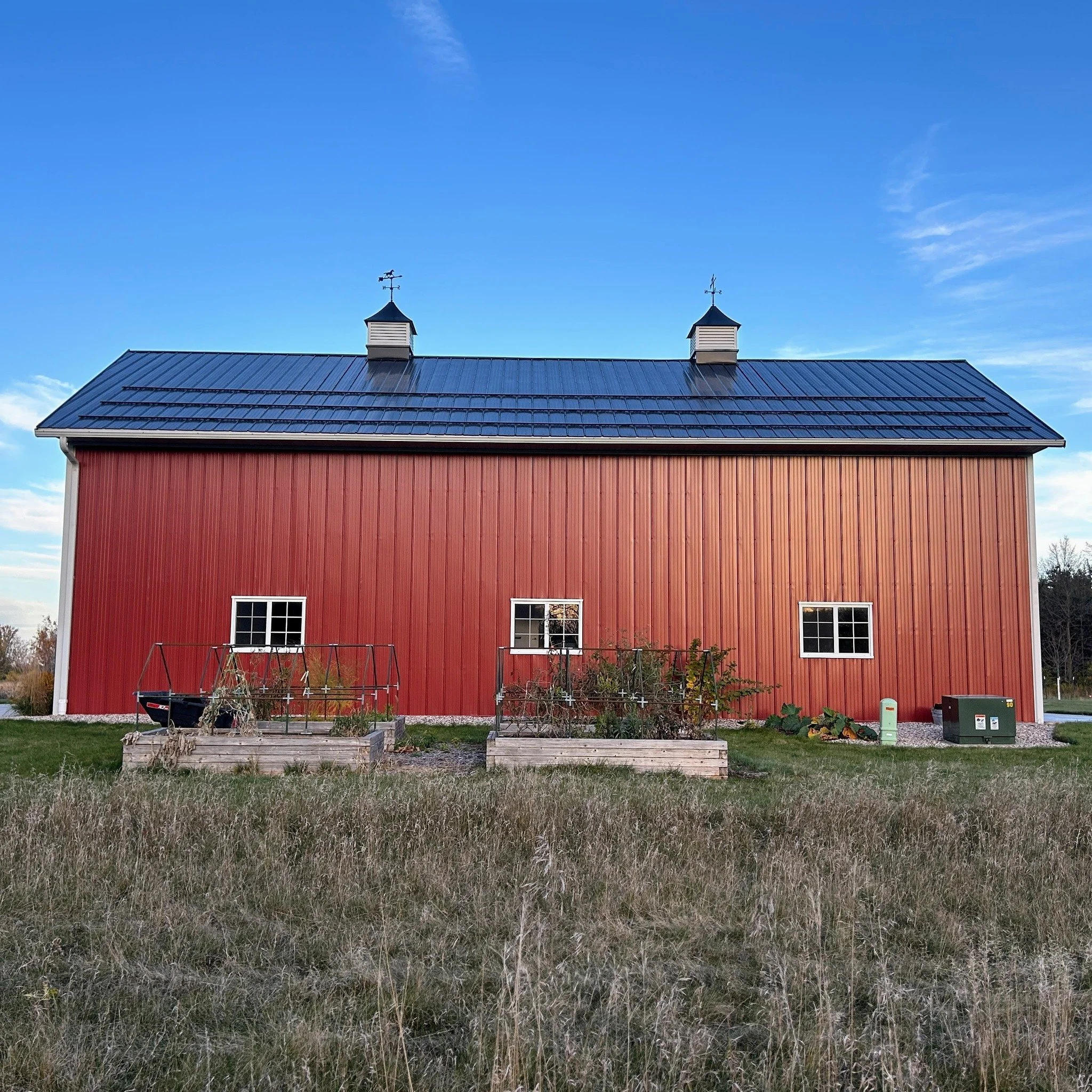 A red barn with a blue metal roof and two small cupolas with weather vanes on top, situated in a grassy area under a clear blue sky.