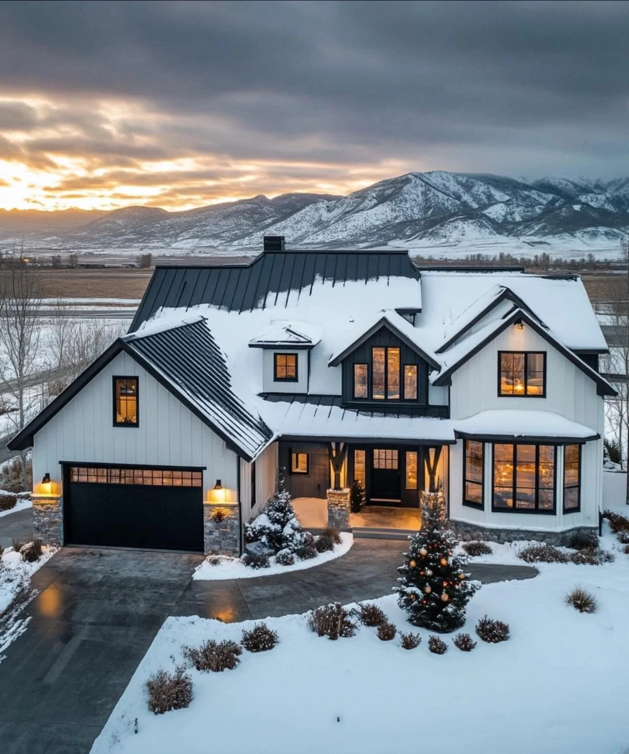 A modern two-story house with black window frames and a black garage door, surrounded by snow, with a Christmas tree outside and mountain scenery in the background at sunset.