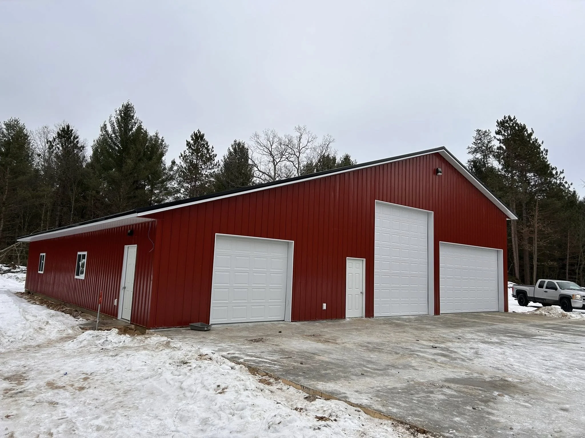 Red metal building with three white garage doors and a side door, situated on snowy ground with a forest background.