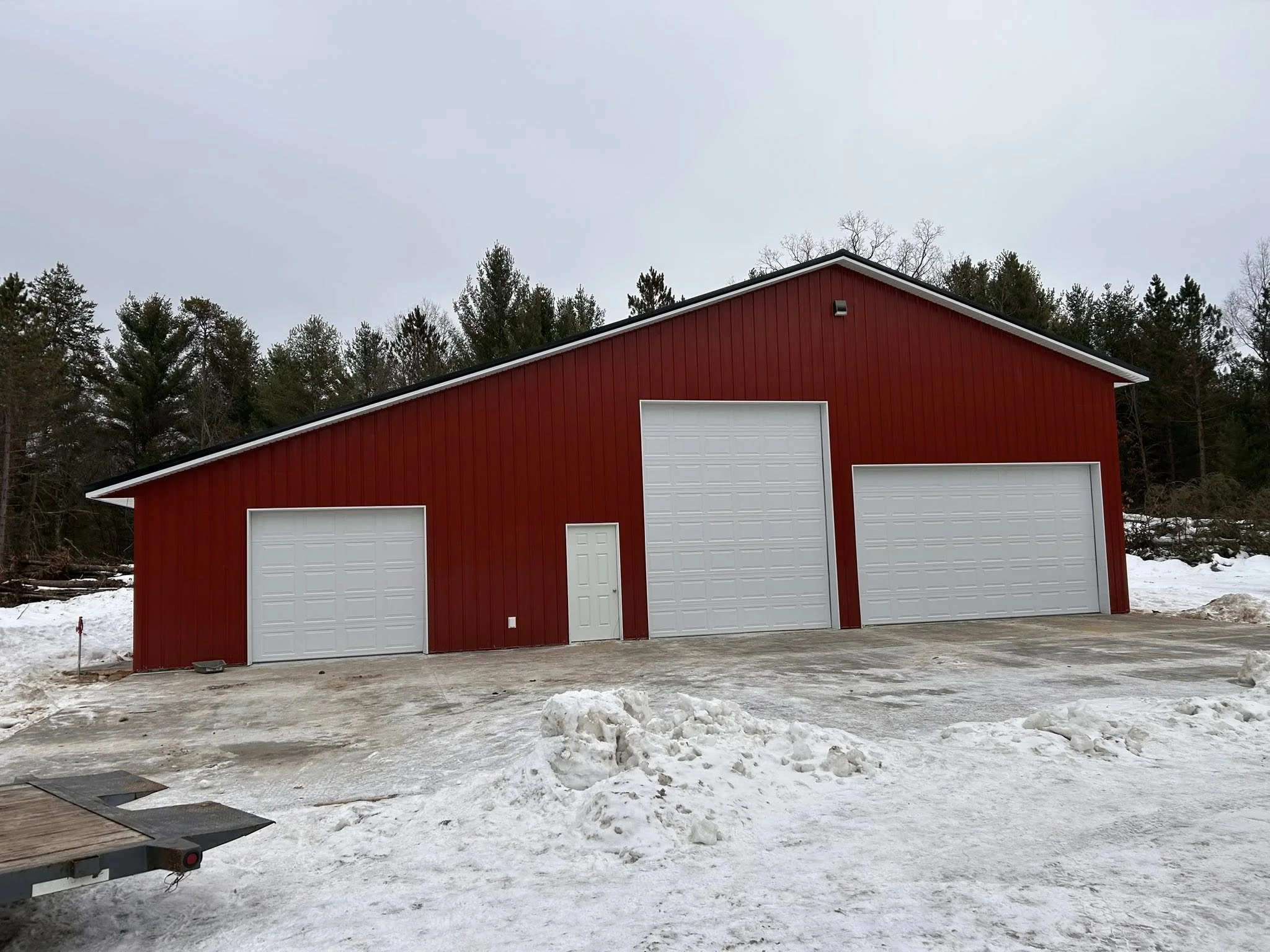 A red metal building with three white roll-up garage doors and a small white pedestrian door in the middle, set in a snowy landscape with trees in the background.